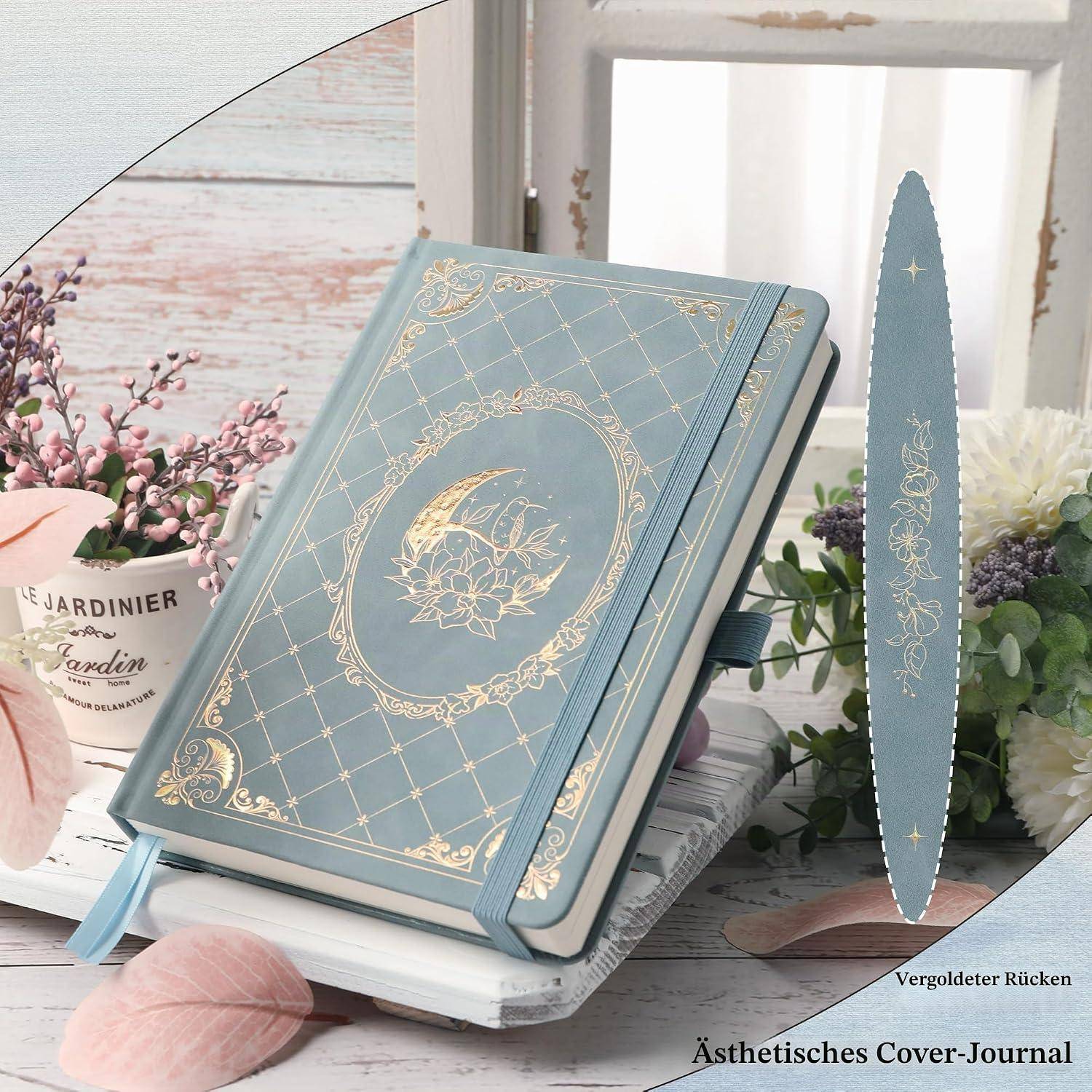 Blau-goldenes Blumenmotiv-Notizbuch mit Elastikband, Bändchen-Lesezeichen und vergoldetem Rücken, präsentiert auf gestapelten Büchern mit Blumen und Fenster-Hintergrund.