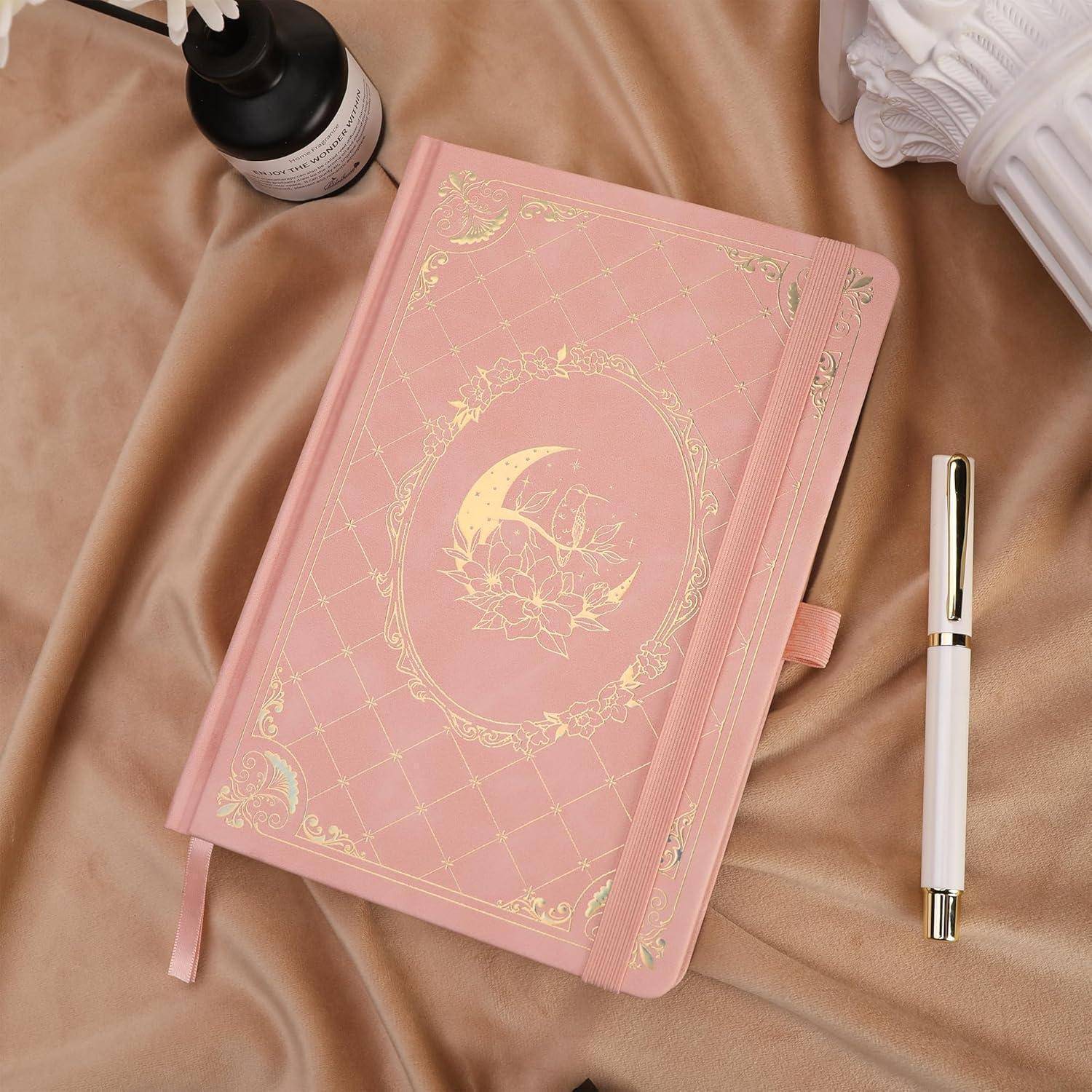 Rosa Hardcover-Notizbuch mit goldenem Halbmond-Design liegt auf Satinstoff neben einem Stift, einer Flasche und einem gefalteten Handtuch.
