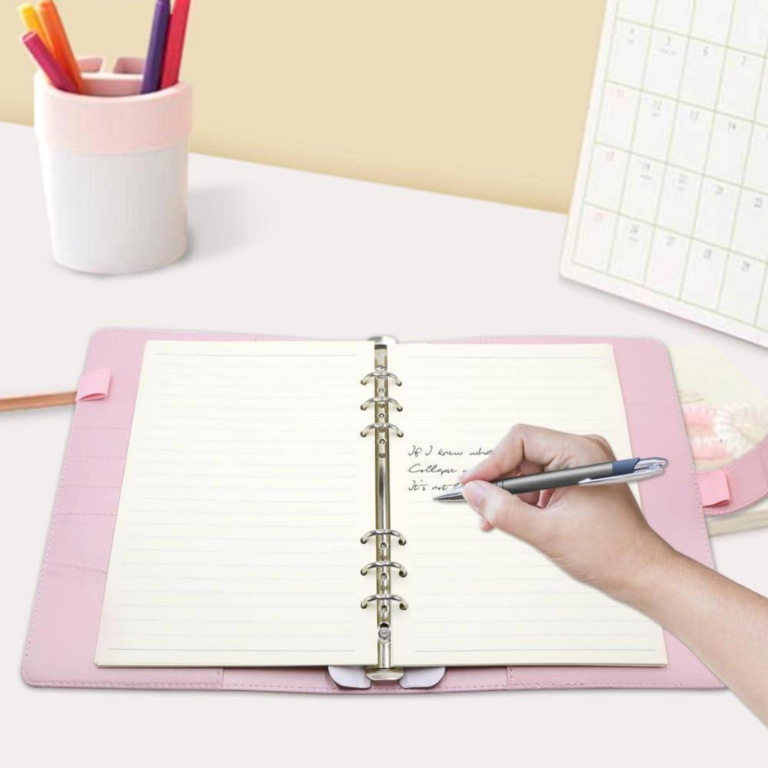 Hand schreibt in einem offenen rosa spiralgebundenen Planer auf einem Schreibtisch, mit Bleistiften, einem Stiftbecher und einem Wandkalender in der Nähe.
