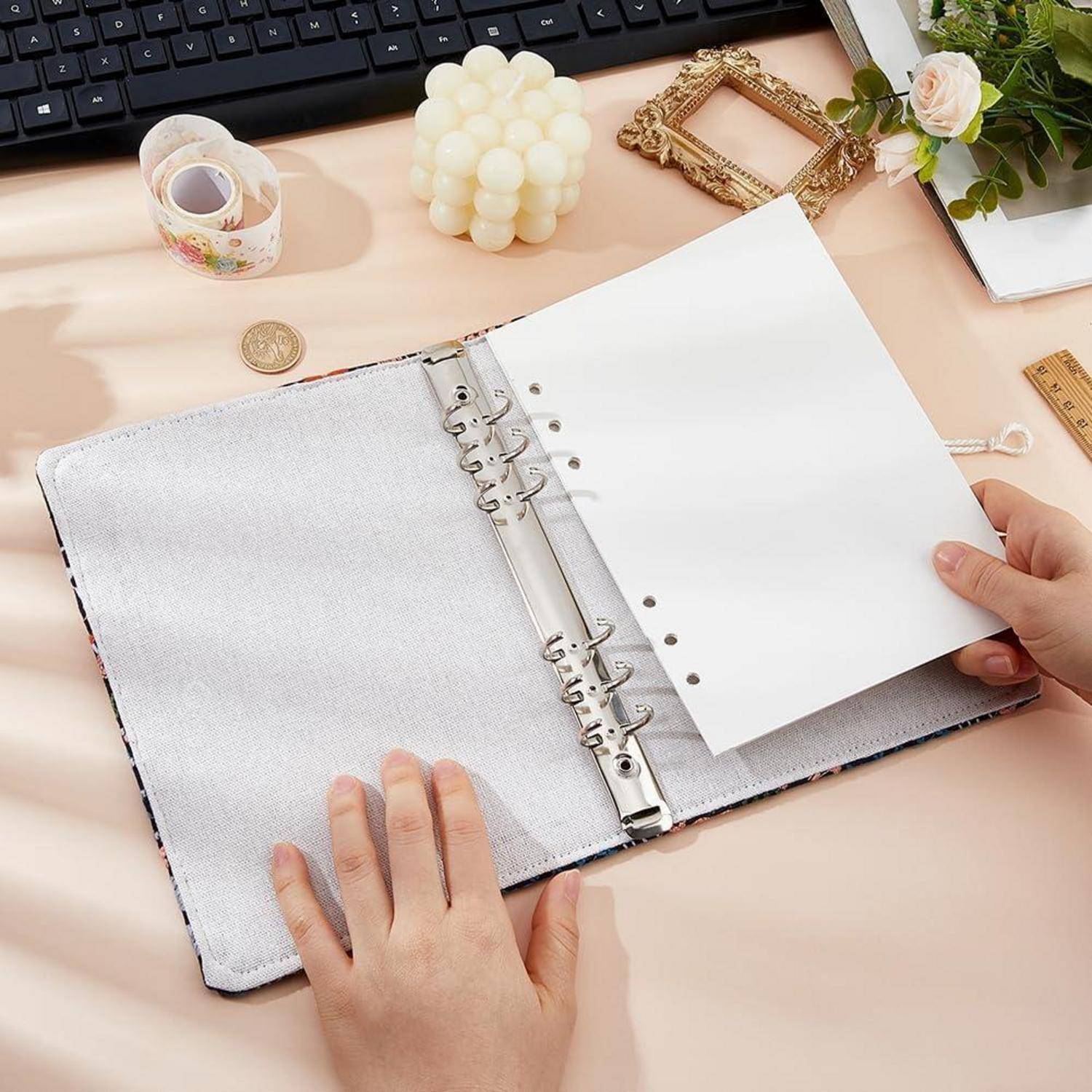 Hände öffnen einen Ringordner mit leerem Papier auf einem Schreibtisch neben einer Tastatur, einer Kerze, Klebeband, einem Rahmen, einer Blume und einem Lineal.