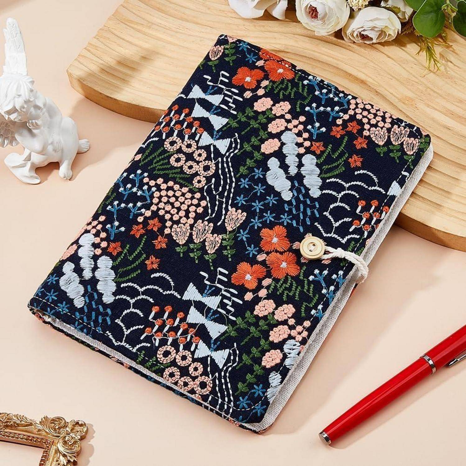 Bestickter Stoffnotizbucheinband mit Blumen- und Kranichmustern, mit Knopfverschluss verschlossen, auf einer Tischplatte mit einem roten Stift und dekorativen Blumen angezeigt.