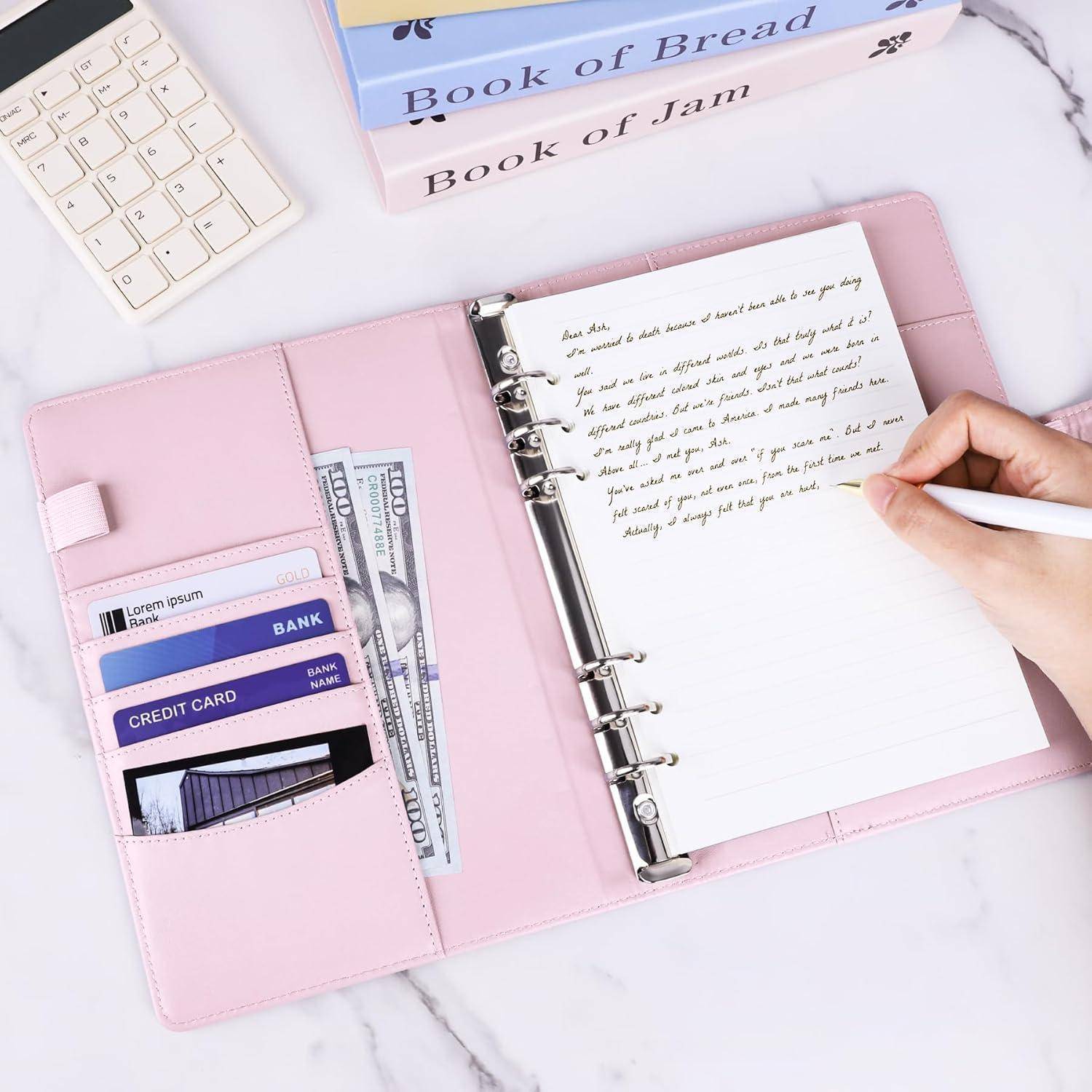 Rosa Ordnerplaner mit handschriftlichen Notizen, eine Hand schreibt auf der Seite, Karten und Bargeld in den Taschen, mit einer Tastatur und Büchern in der Nähe.