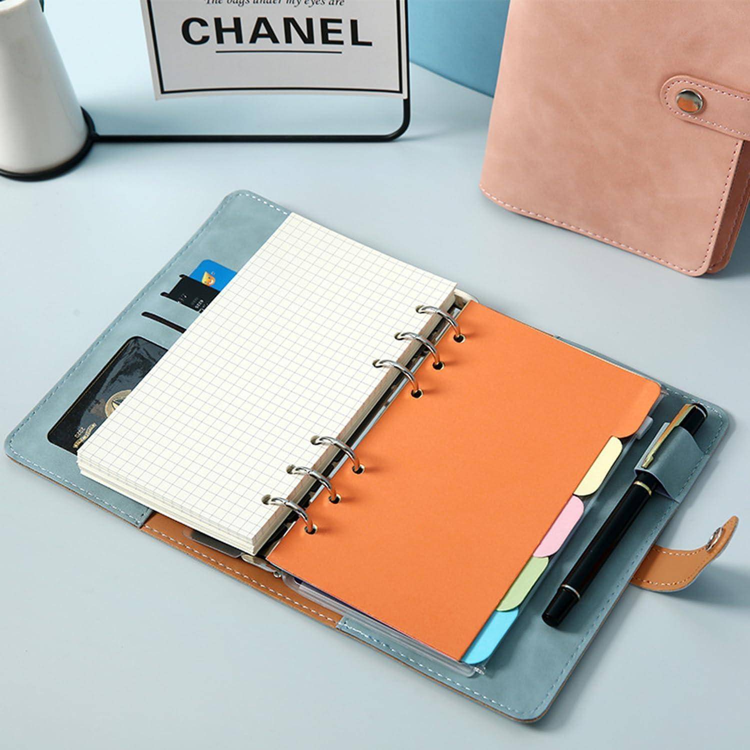Offener Ringordner-Planer mit Rasterpapier, orangenem Trennblatt, Reitern, Karten und Stift auf einem Schreibtisch; eine rosa Tasche sitzt in der Nähe.