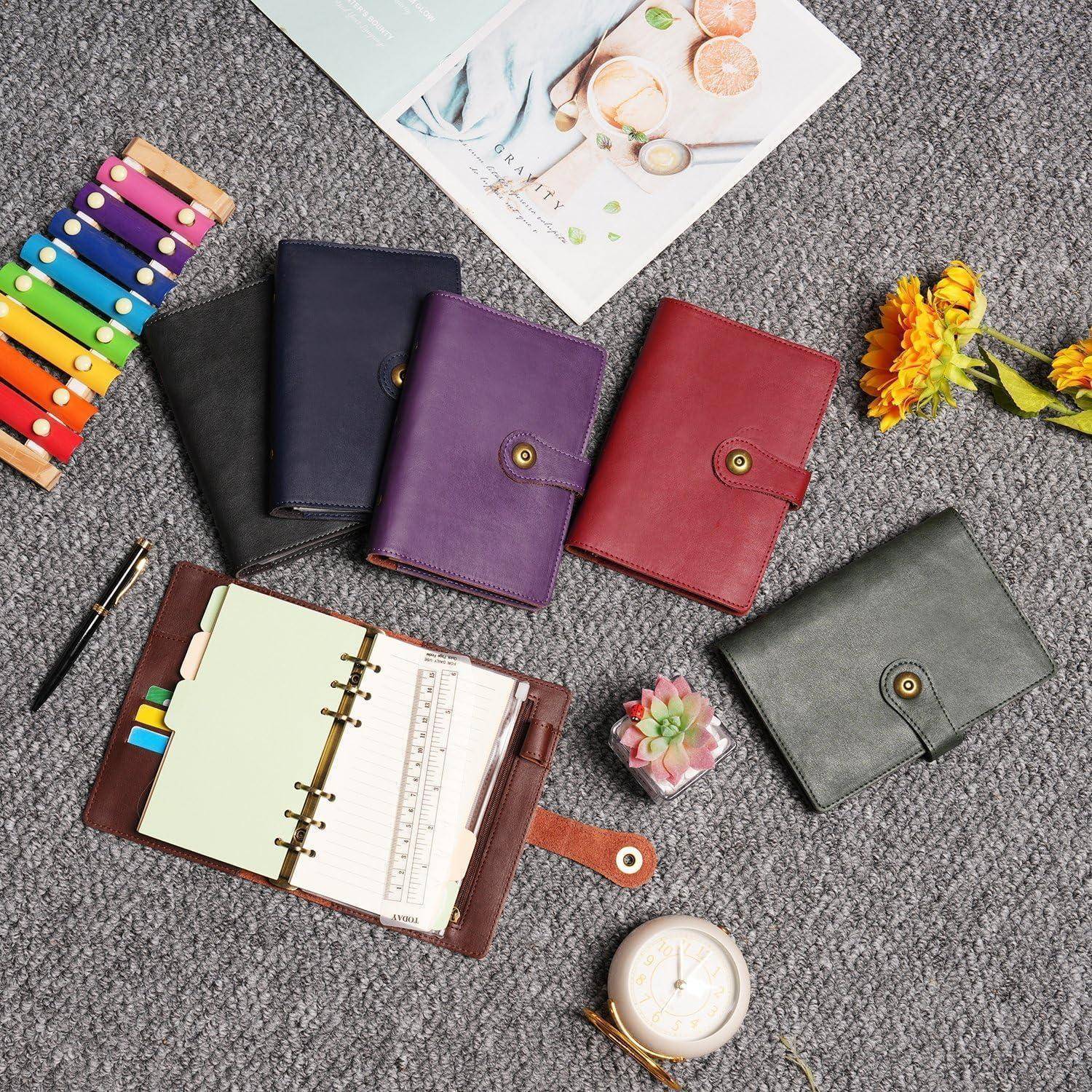 Flache Anordnung von farbenfrohen Lederplanern und einem offenen Ordner mit Reitern auf einem grauen Teppich, mit einem Stift, Blumen und einer kleinen Uhr.