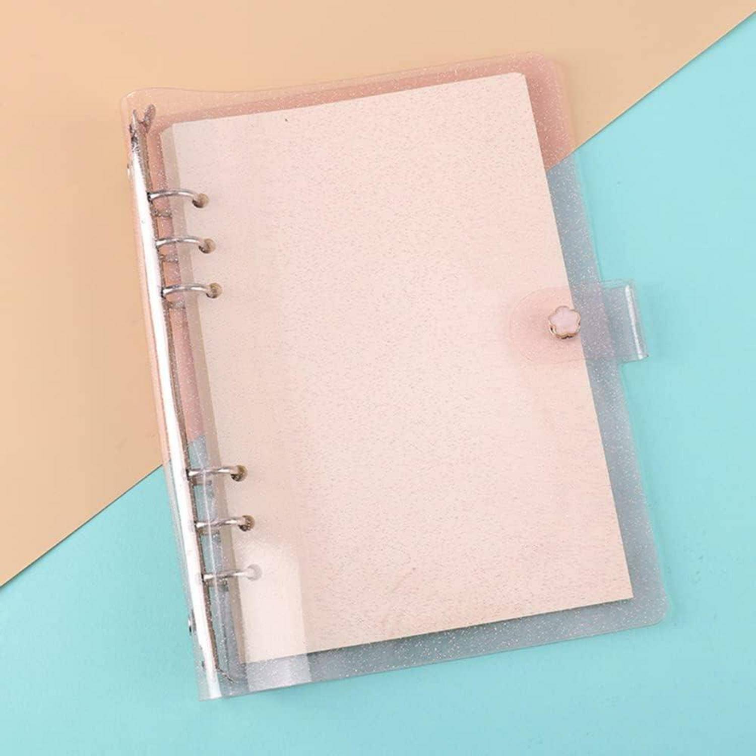 Notizbuch mit transparentem Einband und leeren Seiten sowie Metallringen auf einem pastellfarbenen beige-blauen Hintergrund.