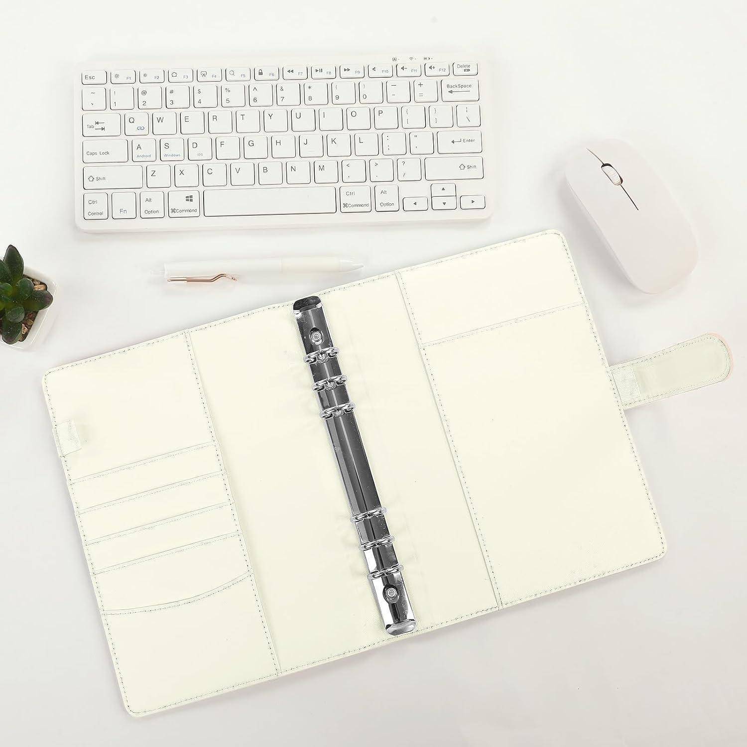 Offener cremefarbener Lederbinder/Planer mit Sechslochbohrung neben einer weißen Tastatur und Maus auf einem sauberen Schreibtisch.