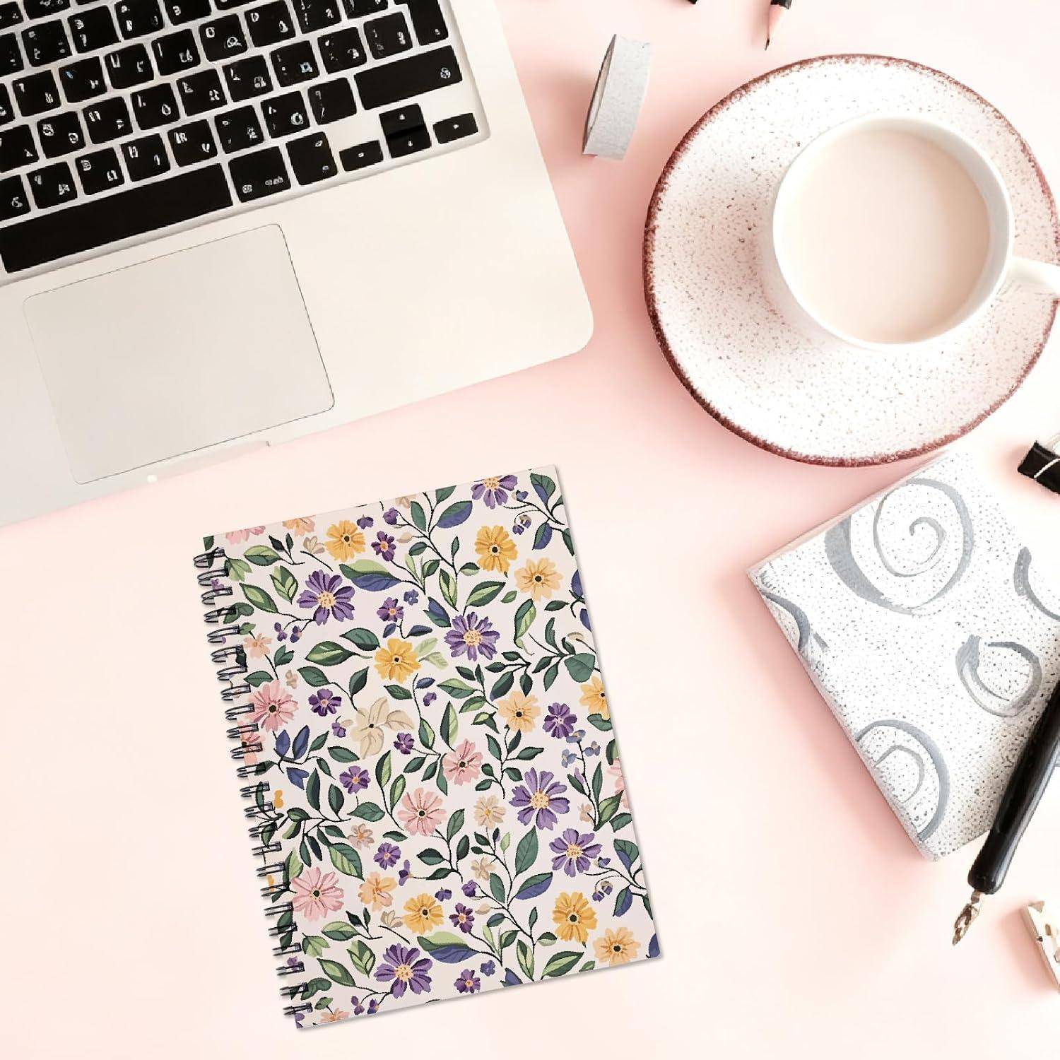 Flachaufnahme eines Laptops, einer Kaffeetasse, eines Notizbuchs mit Blumenmuster in Spiralbindung, eines Stifts und eines klein zusammengefalteten Papiers auf einem pastellrosa Schreibtisch.