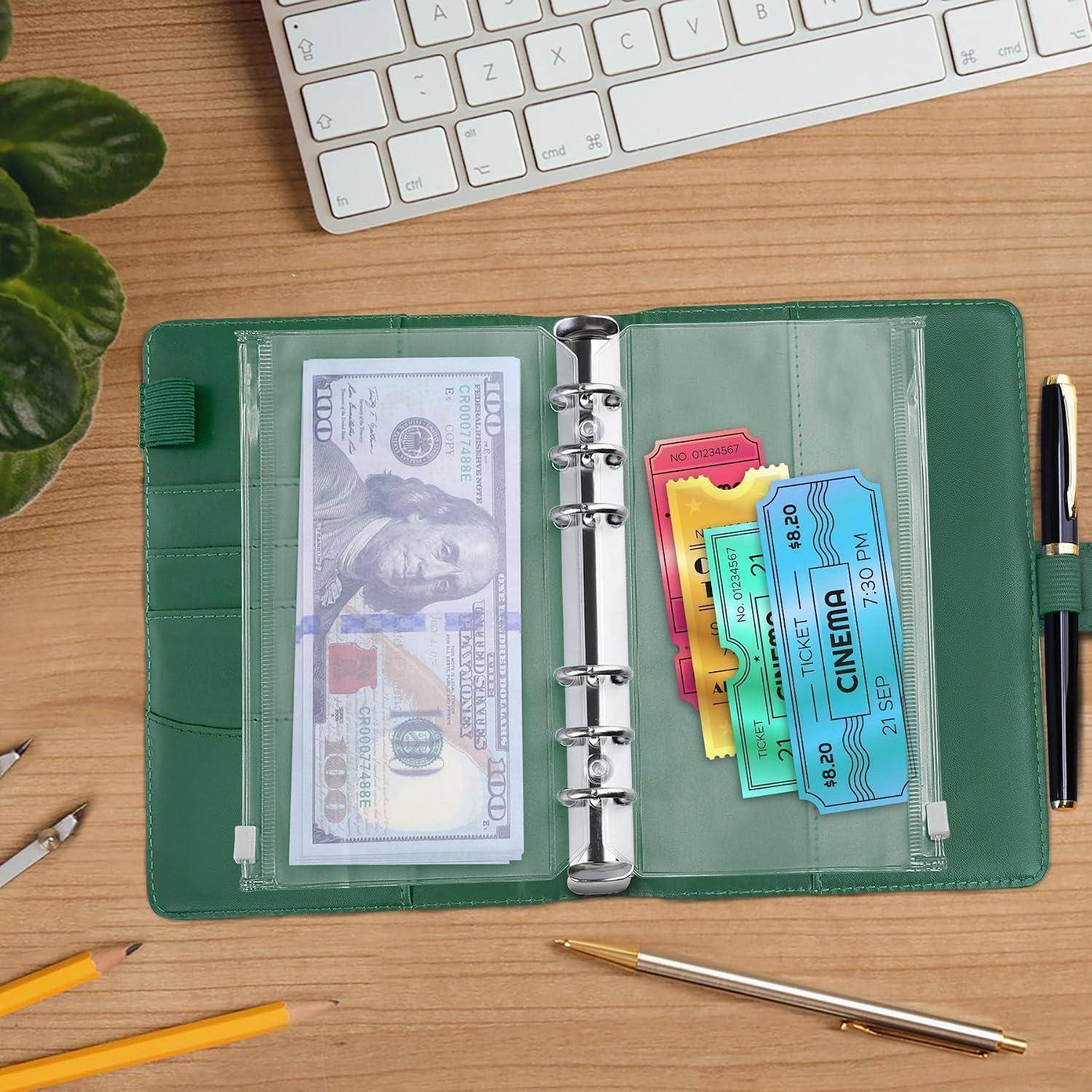 Ein offener grüner Ordner auf einem Schreibtisch enthält einen Geldschein und bunte Kinokarten in durchsichtigen Taschen, mit einer Tastatur und Stiften in der Nähe.