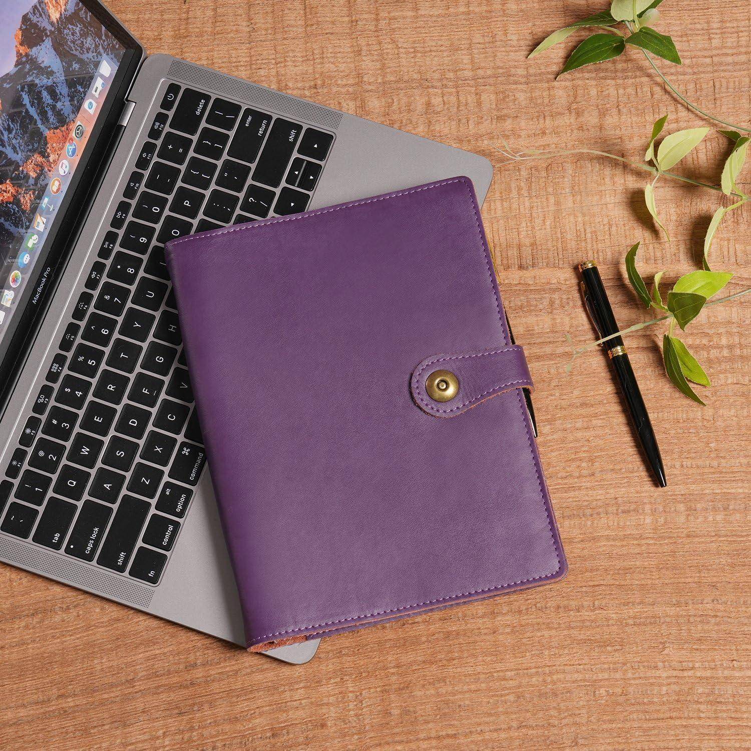 Violettes Notizbuch auf einem Laptop neben einem Stift und einer kleinen Pflanze auf einem Holzschreibtisch.