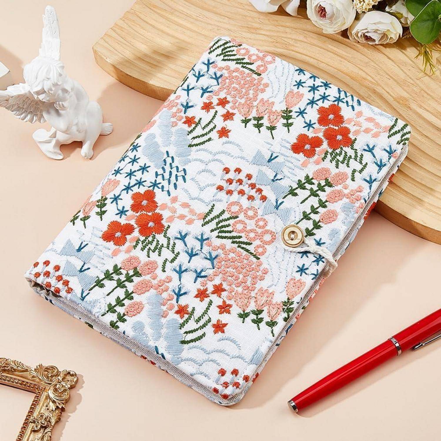 Notizbuch oder Planer aus Stoff mit Blumenmuster mit einer Goldknopfschließe auf einem Pastell-Schreibtisch, neben einem roten Stift und dekorativen Gegenständen.