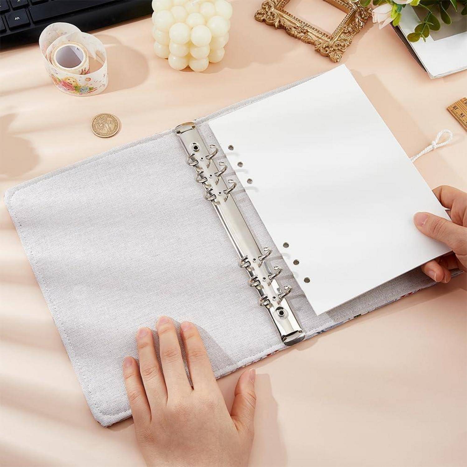 Hände öffnen ein weißes Notizbuch mit sechs Ringen auf einem Pastell-Schreibtisch mit einer Schlaufе für Stifte, leeren Seiten und Schreibwaren in der Nähe.