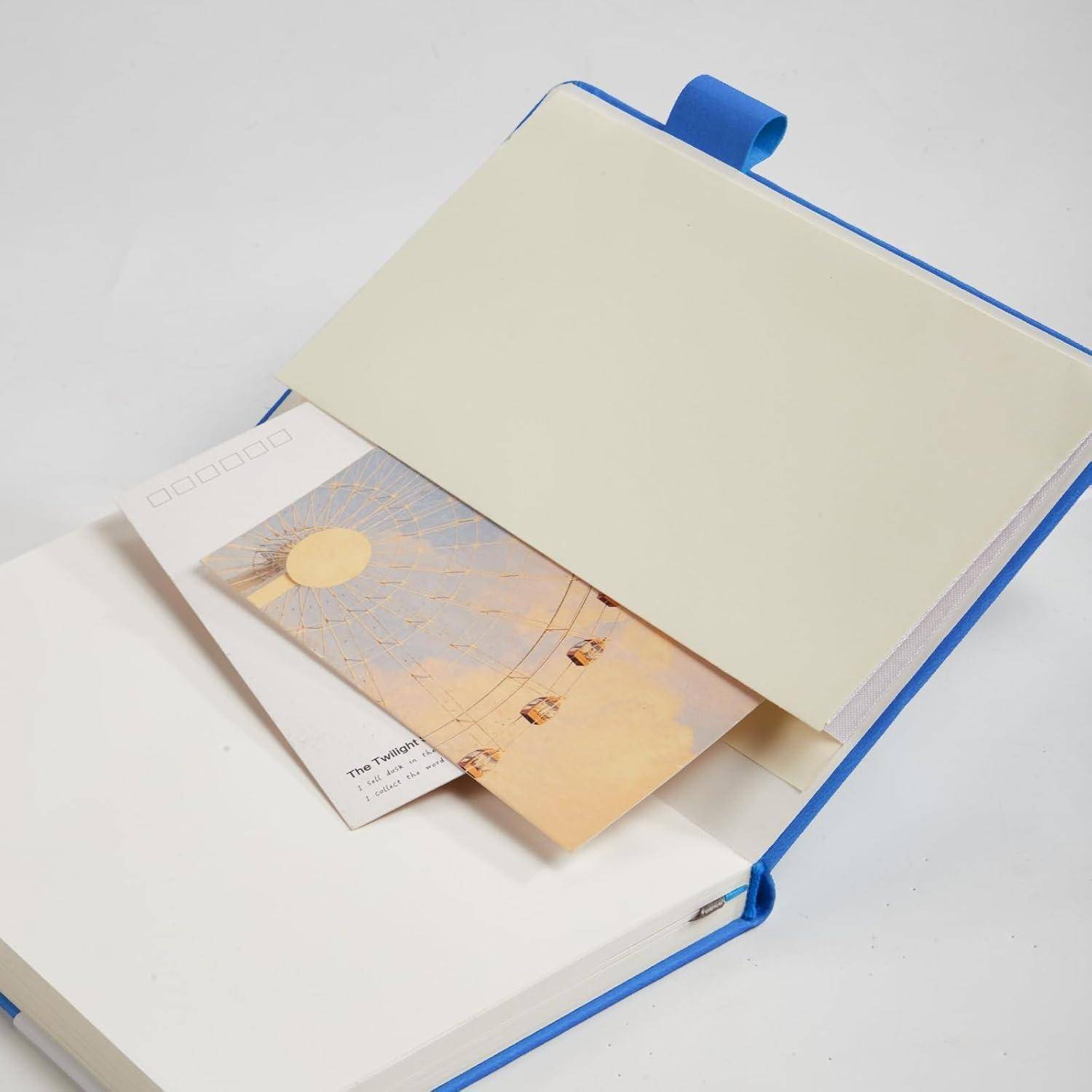 Aufgeschlagenes blaugebundenes Notizbuch oder Planer mit cremefarbener Umschlagklappe, in dem teilweise eine Postkarte oder ein Fotodruck eingesteckt ist.