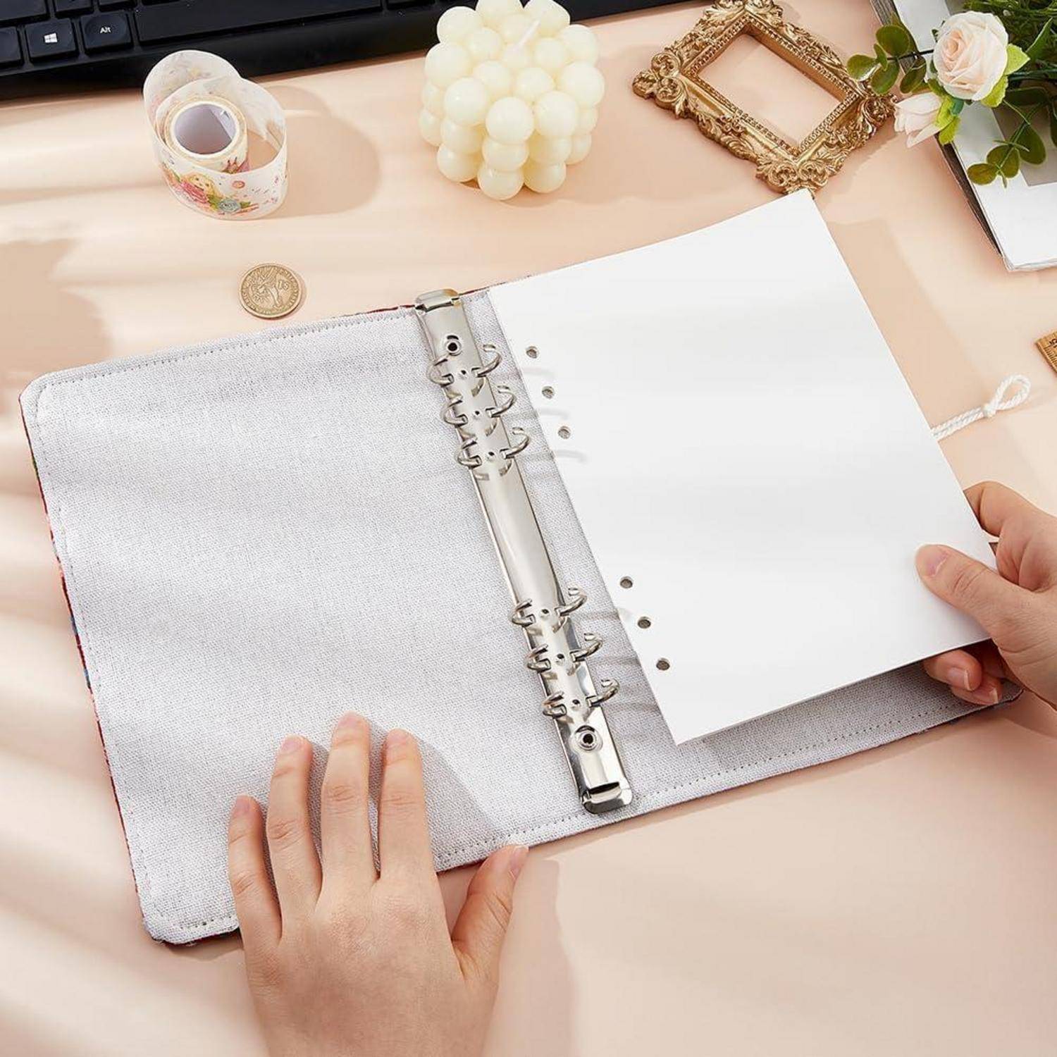 Hände, die einen weißen Ringbinder auf einem Pastellschreibtisch mit Washi-Tape, Kerze und dekorativem Rahmen in der Nähe öffnen.