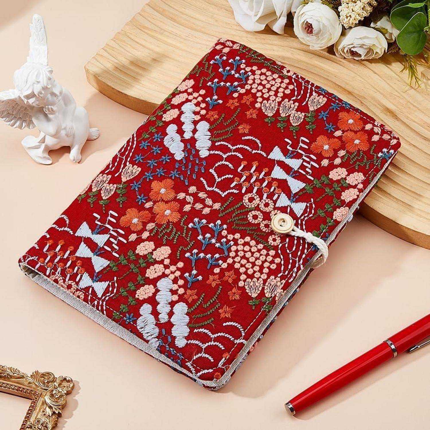 Rotes, blumig besticktes Notizbuchcover mit Knopfverschluss, gezeigt mit einem roten Stift und kleinen dekorativen Figuren auf einem pastellfarbenen Schreibtisch.
