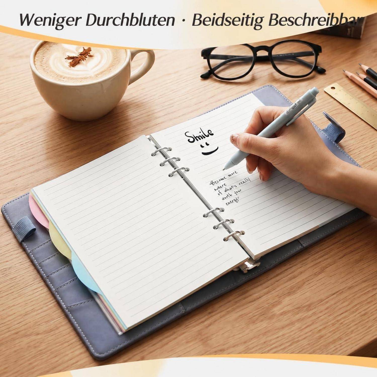 # Hand schreibt in einem Ringbuch neben Kaffee und Brille auf einem Schreibtisch; Der deutsche Text oben sagt „Weniger Durchbluten · Beidseitig Beschreibbar