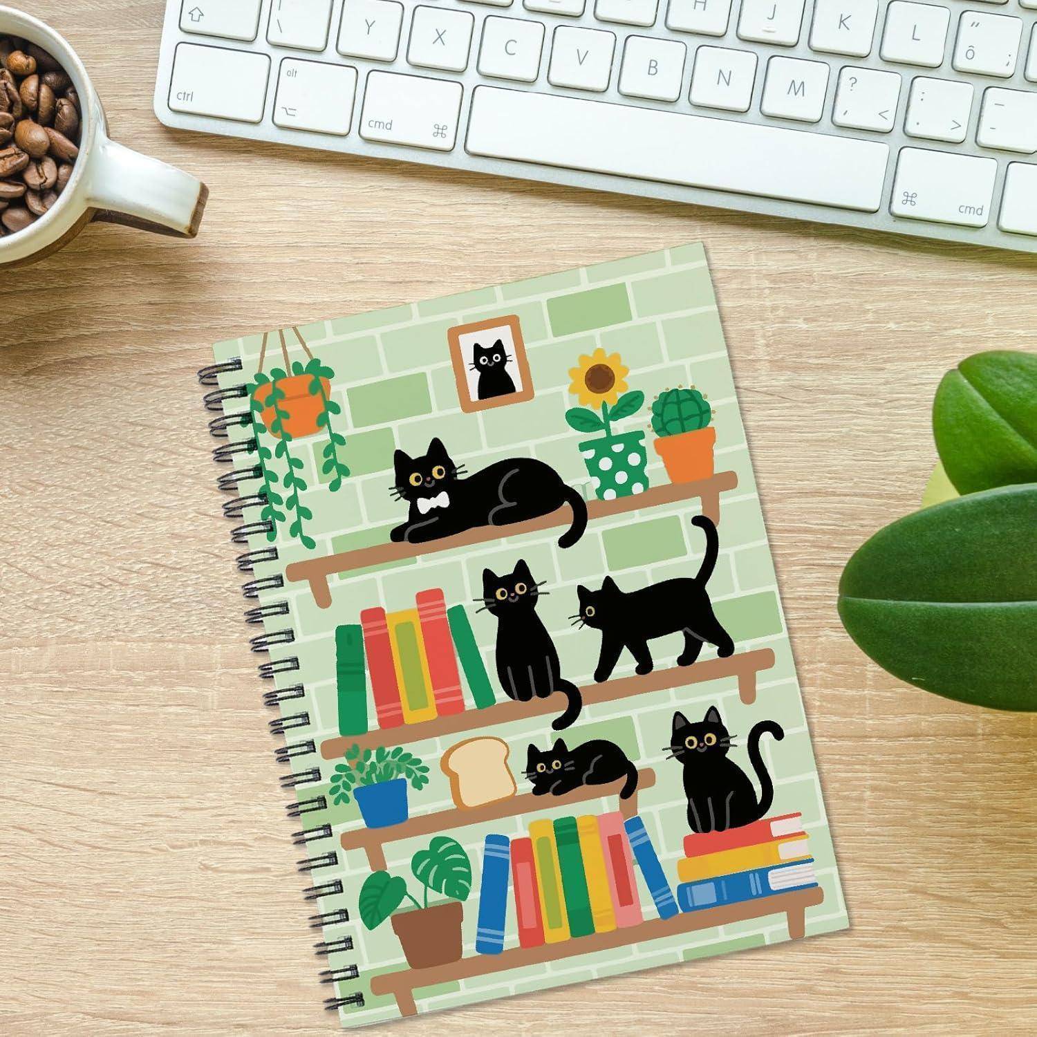 Ein Spiralnotizbuch mit grünem Backsteinmuster-Umschlag, das schwarze Katzen auf Regalen zwischen Büchern und Topfpflanzen zeigt, auf einem Schreibtisch neben einer Tastatur und einer Kaffeetasse.