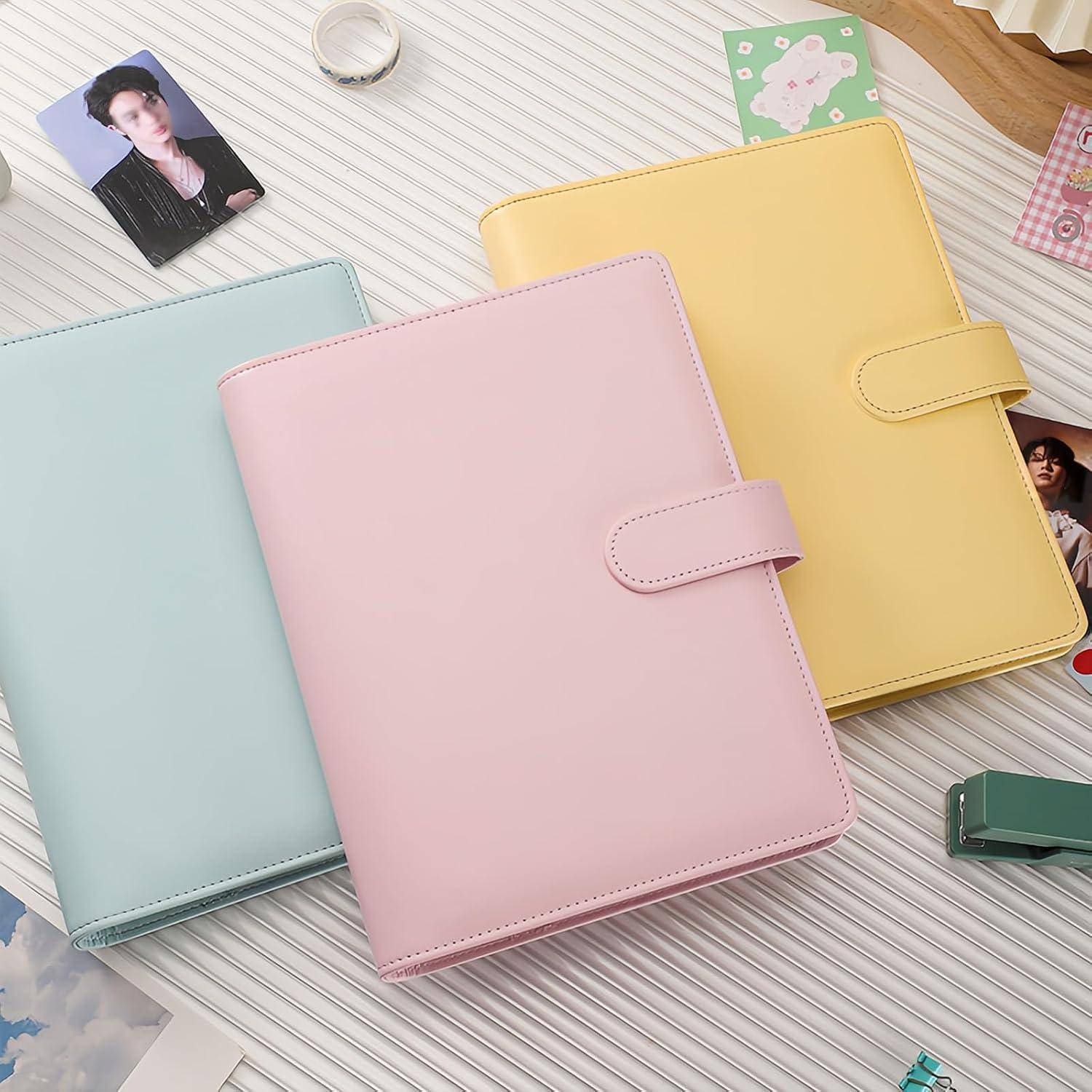 Drei Ringbuch-Planer in Pastellfarben – Minzgrün, Rosa und Gelb – sind auf einem gestreiften Schreibtisch mit einigen kleinen Fotokarten angeordnet.