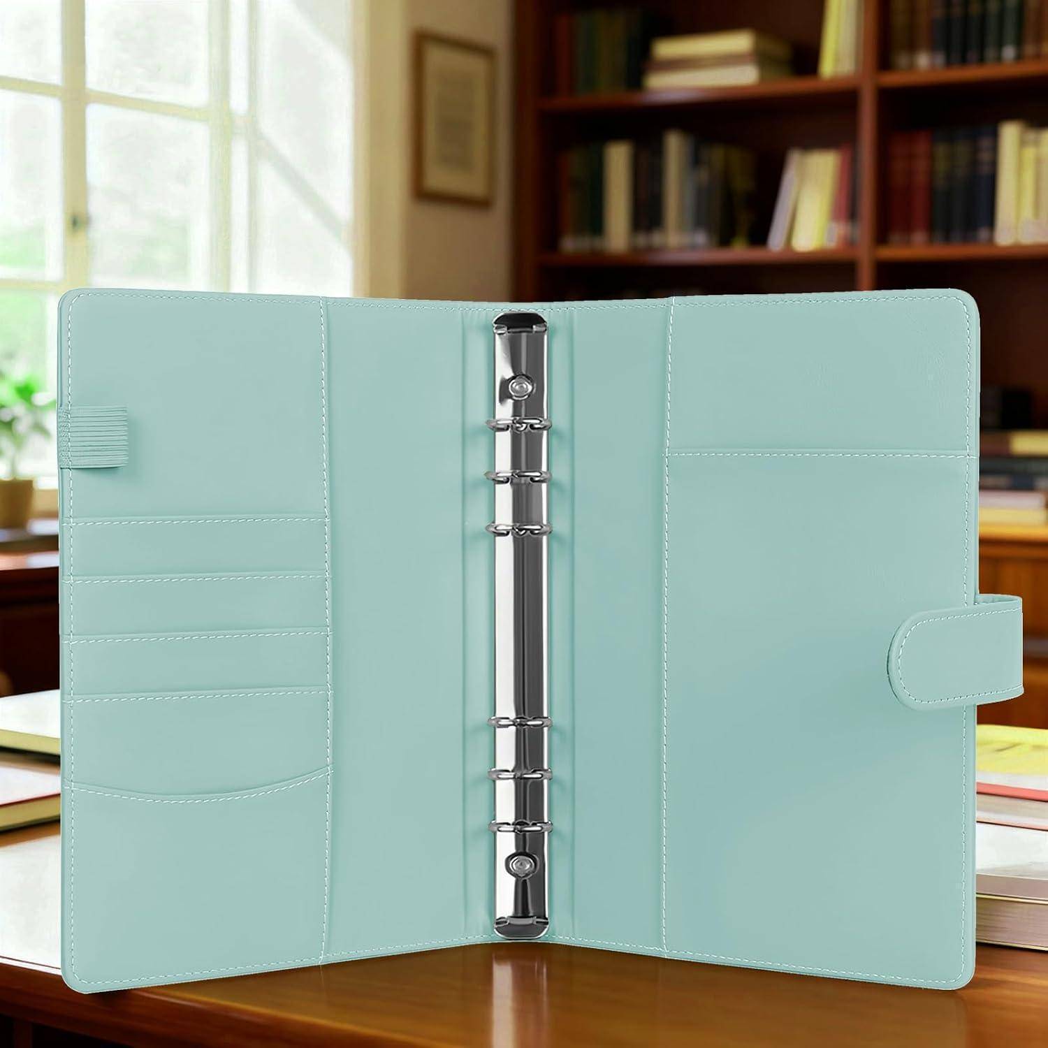 Offener mintgrüner Ringordner-Notizbuch auf einem Holzschreibtisch, mit Taschen im Inneren und Bücherregalen im Hintergrund.