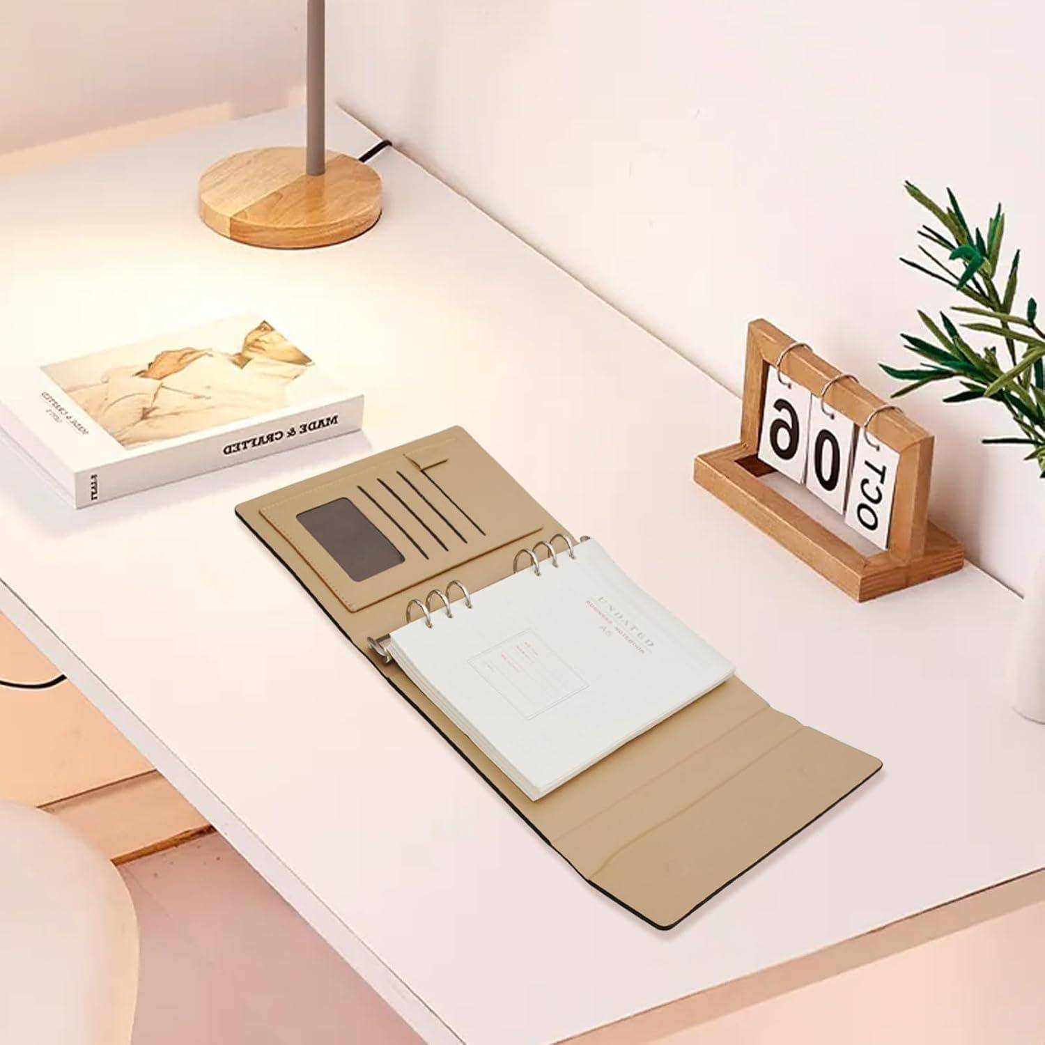 Minimales Schreibtischset-up mit einem offenen beigen Spiralnotizblock auf einem weißen Tisch, einer Holz-Flip-Uhr, die 00:30 Uhr anzeigt, einem Buch, einer Lampensockel und einer Pflanze.