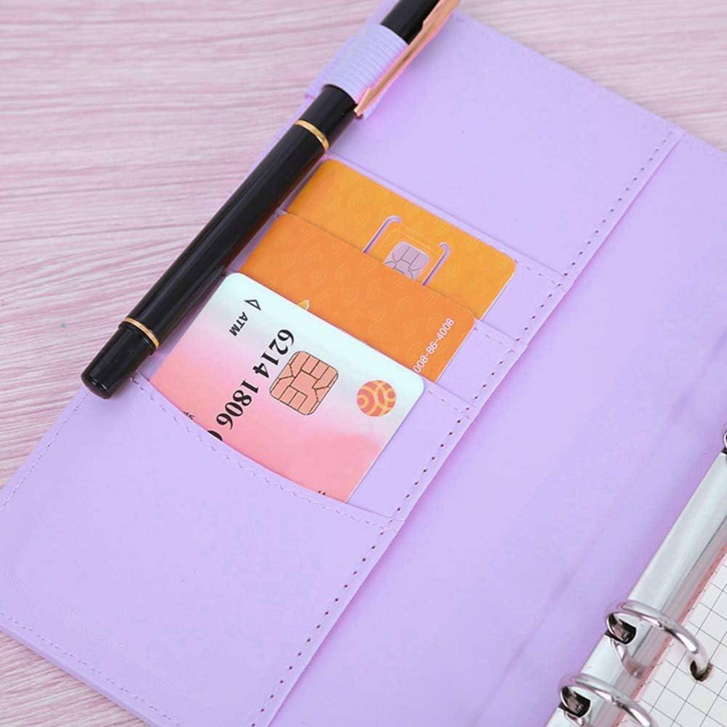 Lavendelfarbener Planer mit Stift und Binderclip offen; Kreditkarte und orangefarbene Karte in Taschen auf einem Schreibtisch.