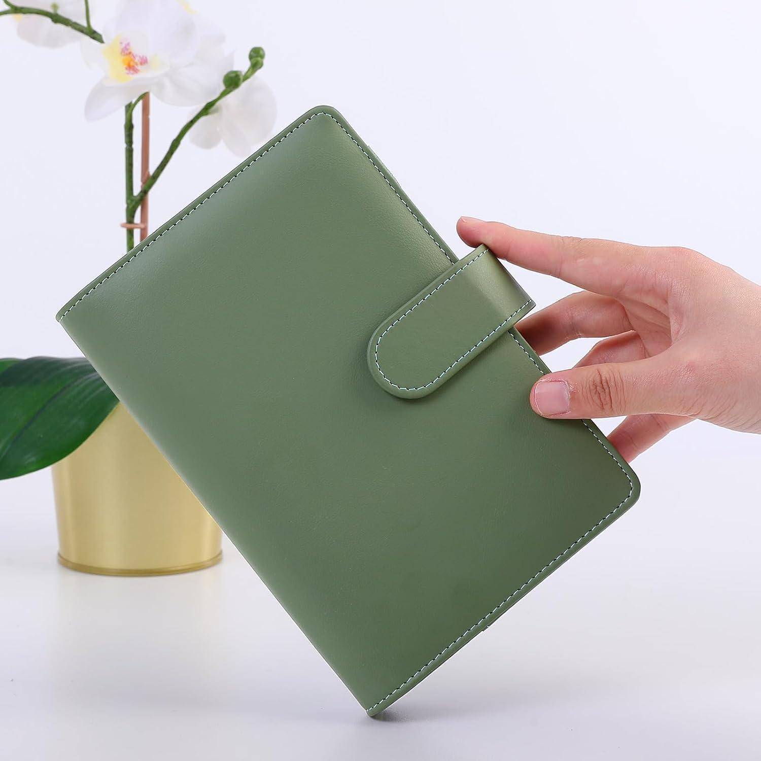 Hand hält einen grünen Lederplaner oder ein Notizbuch mit Riemenverschluss, präsentiert vor einer weißen Orchidee und einer goldenen Vase.