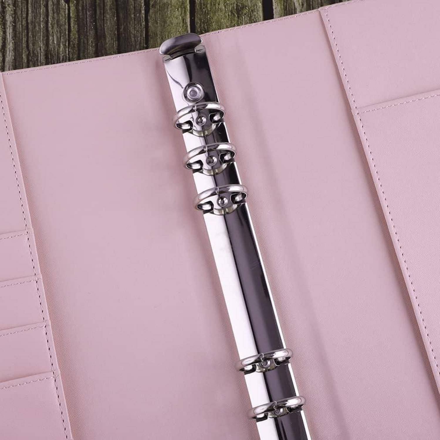 Nahaufnahme eines rosa Ringbindernotizbuchs mit glänzendem silbernem Dreiringmechanismus, der auf einer strukturierten Holzoberfläche geöffnet ist.