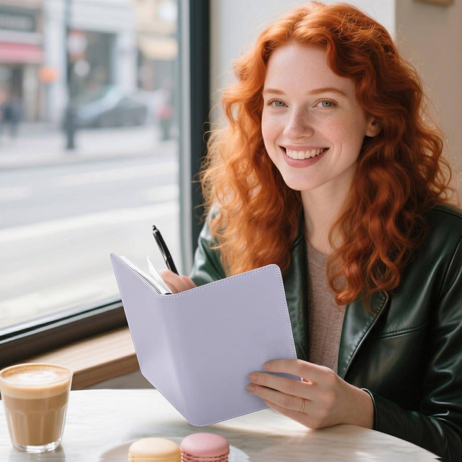 Lächelnde rothaarige Frau in einer grünen Jacke schreibt in ein Notizbuch an einem Cafétisch neben einem Latte neben einem Fenster.