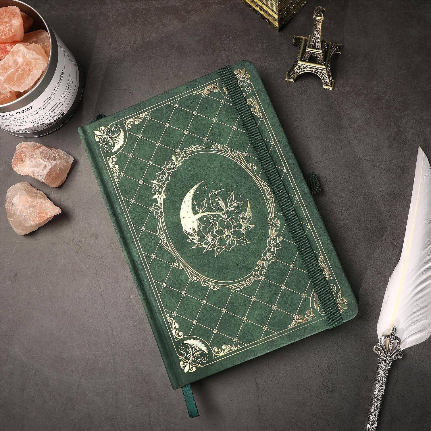Grünes Hardcover-Tagebuch mit Mond- und Blumenmotivenauf dem Einband, umgeben von Kristallen, einer kleinen Eiffelturm-Figur und einer Federfüller.