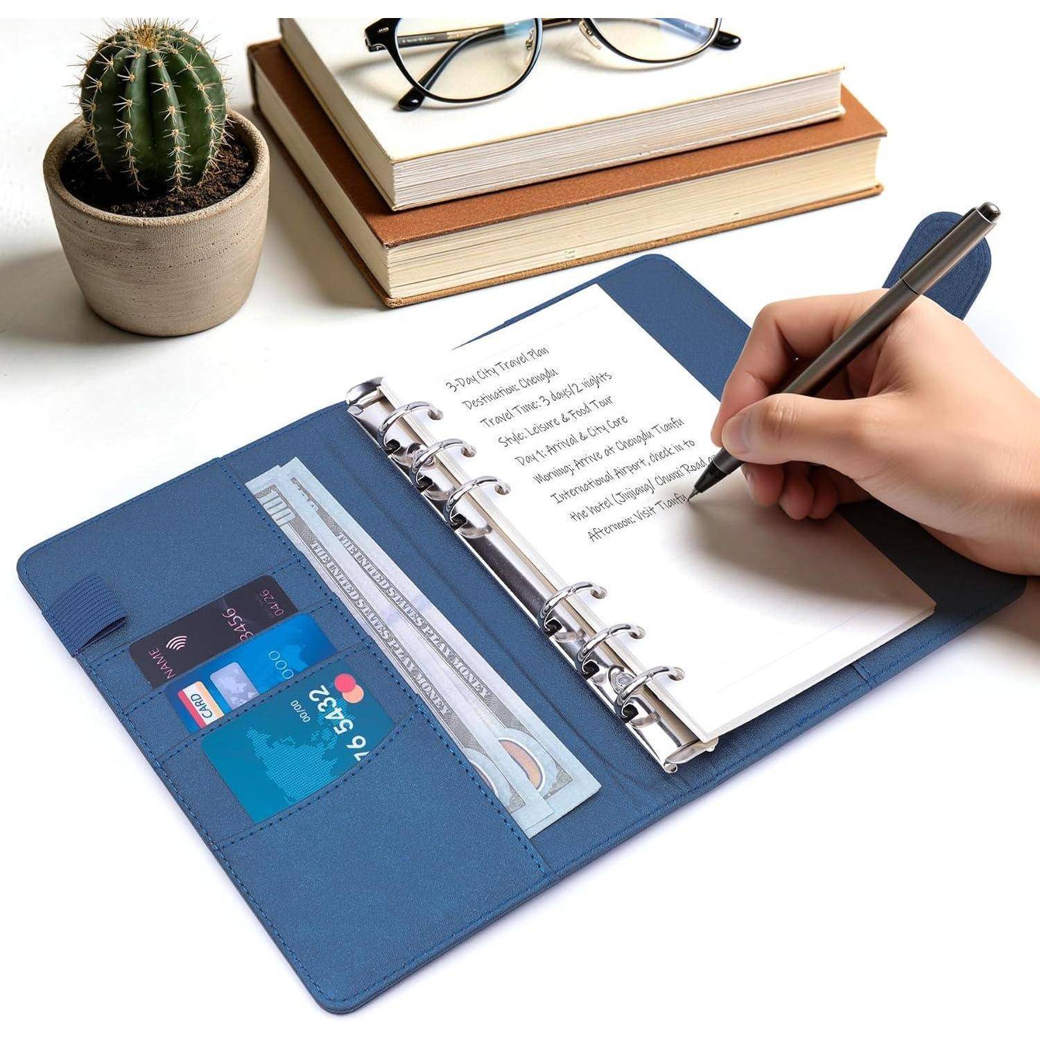 Hand schreibt in einem blauen spiralgebundenen Organizer neben einem Stapel Bücher, einer Brille und einem kleinen Kaktus.