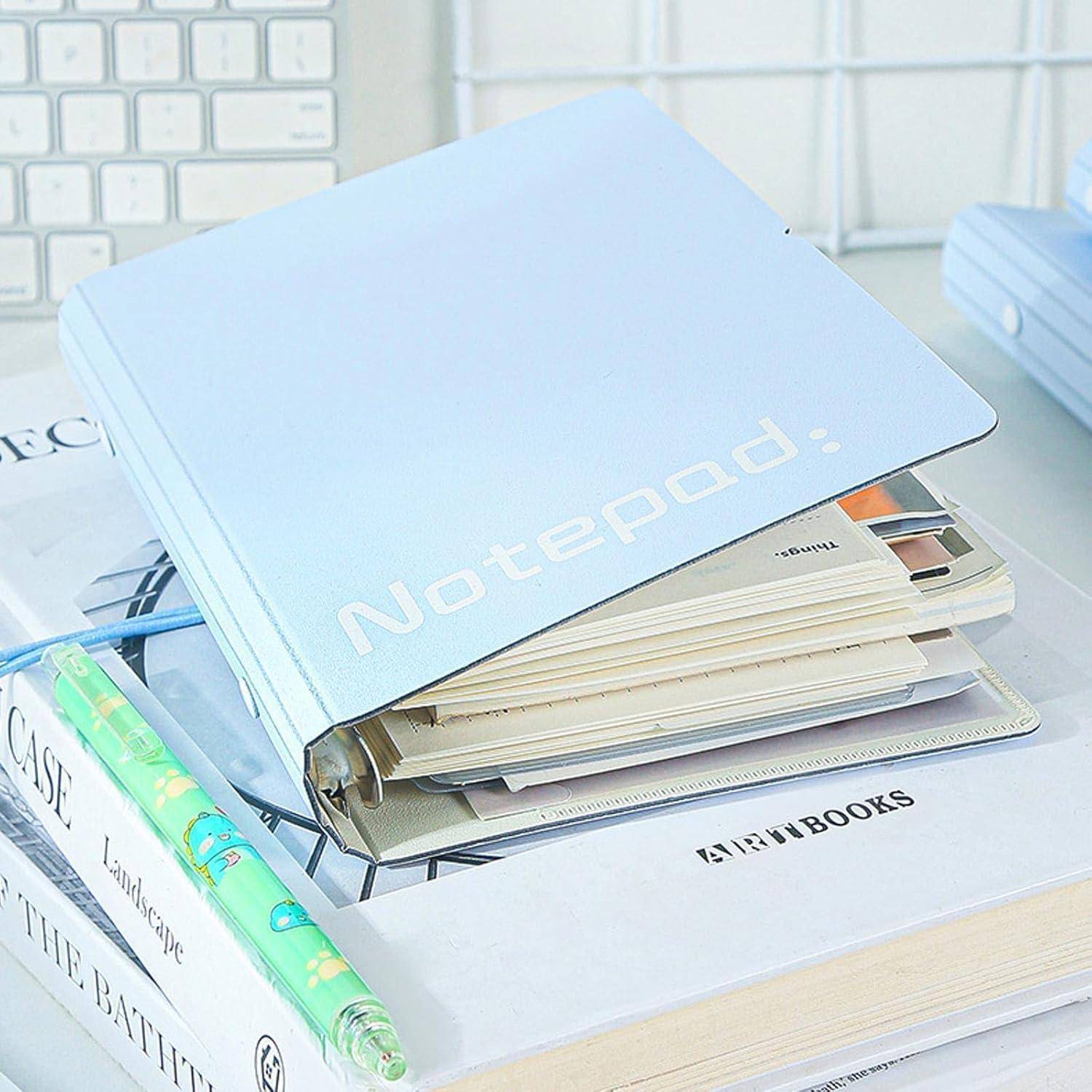 Ein blaues Notizbuch ruht auf einem Stapel von Büchern und Papieren neben einem Bleistift und einer Tastatur auf einem Schreibtisch.
