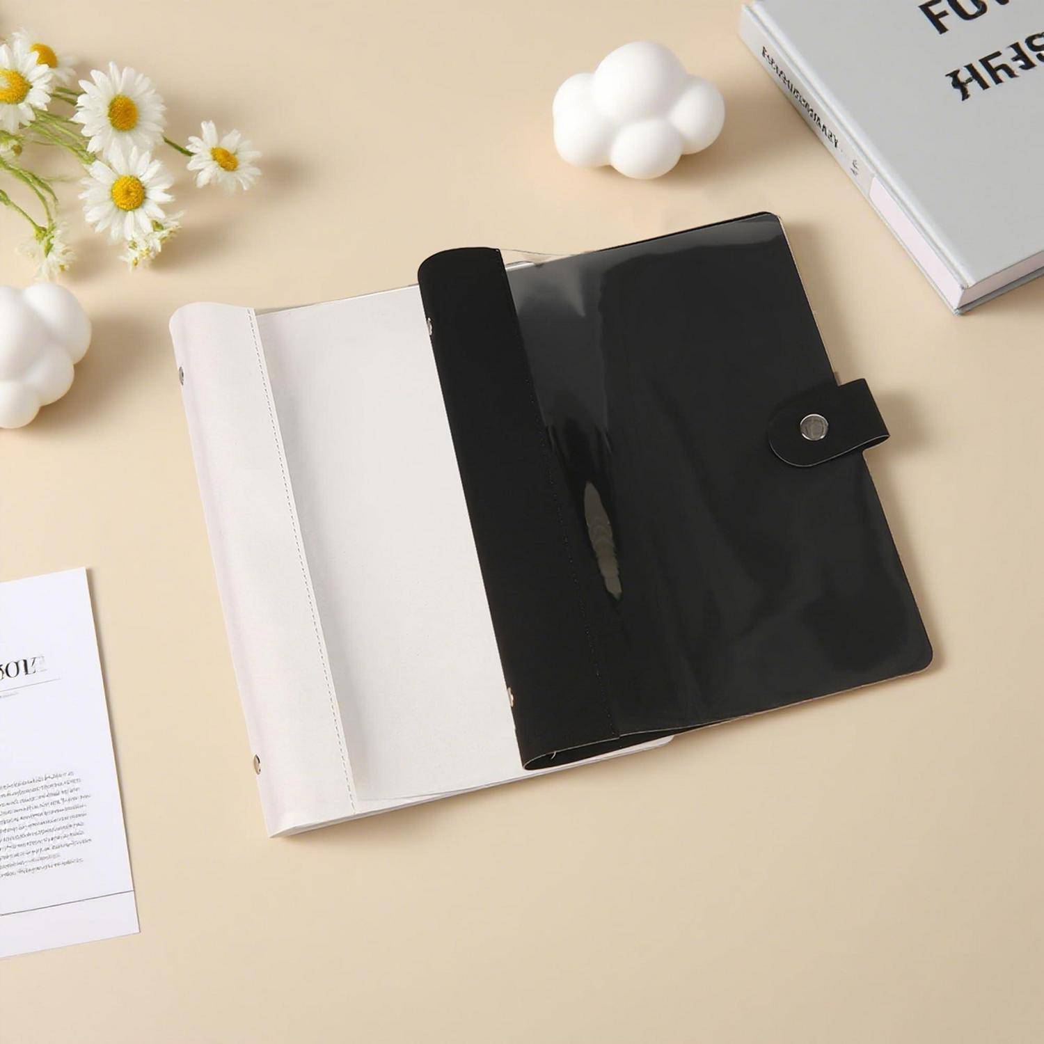 Offener schwarz-weißer Dokumentenordner mit Druckknopfverschluss auf einem beigen Schreibtisch, umgeben von Papieren, einem Buch und kleinen Blumen.