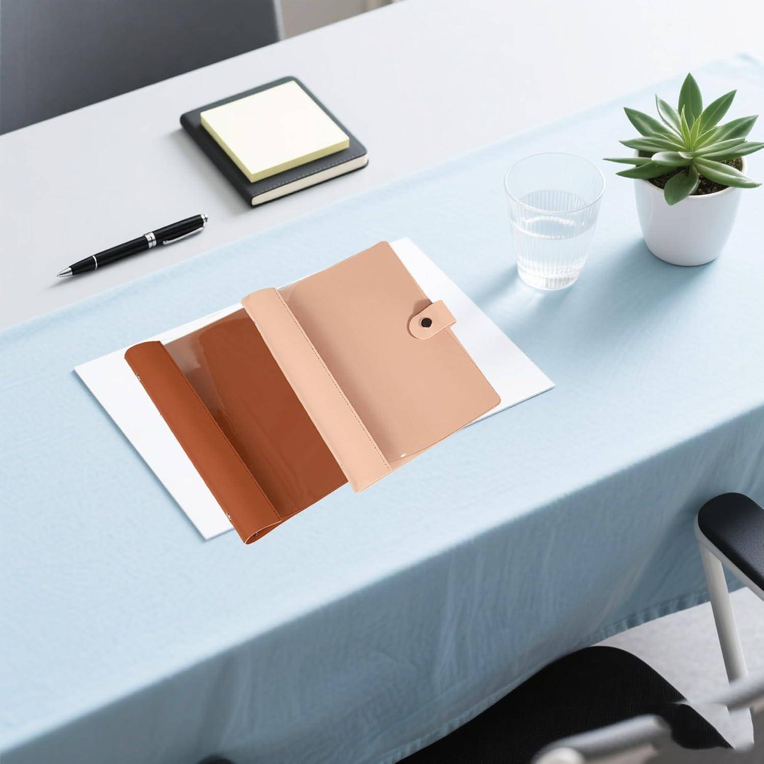 Offener hellbraun-ledergebundener Planer oder Folio auf einem hellblauen Schreibtisch neben einem Stift, Notizbuch, Glas Wasser und einer kleinen Topfpflanze.