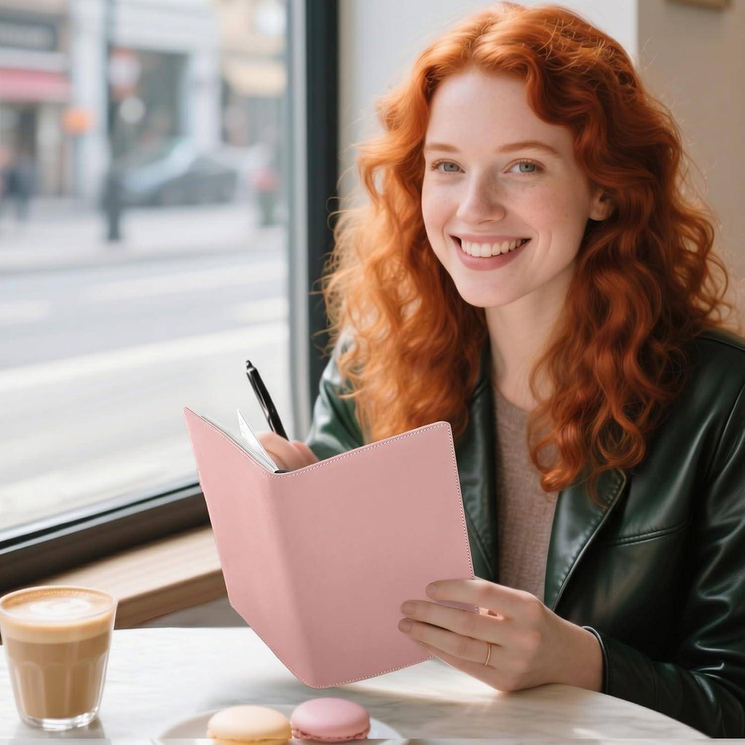 Lächelnde rothaarige Frau in einem Café schreibt in einem rosa Notizbuch neben einem Latte am Fenster.