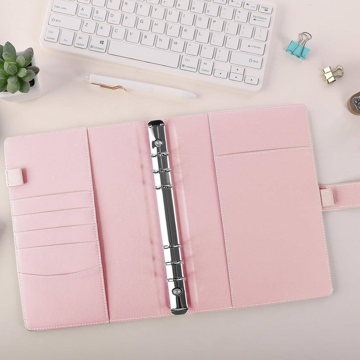 Offener rosa Ringordner-Planer mit Kartensteckplätzen auf einem Schreibtisch neben einer Tastatur, einem Stift, einer Pflanze und Aktenklammern.
