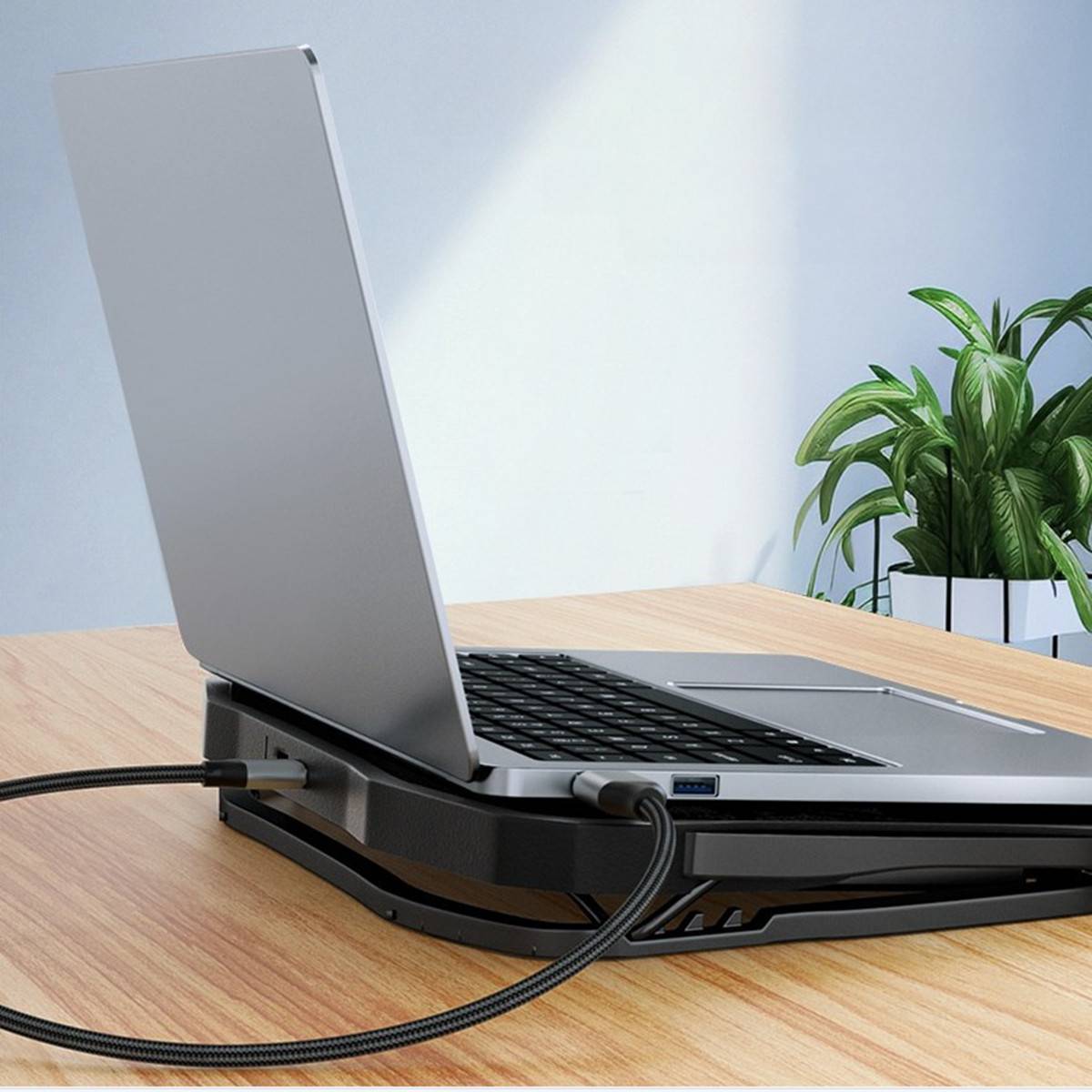 Silberner Laptop auf einem schwarzen Dockingständer mit verbundenen Kabeln, auf einem Holzschreibtisch neben einer Topfpflanze.