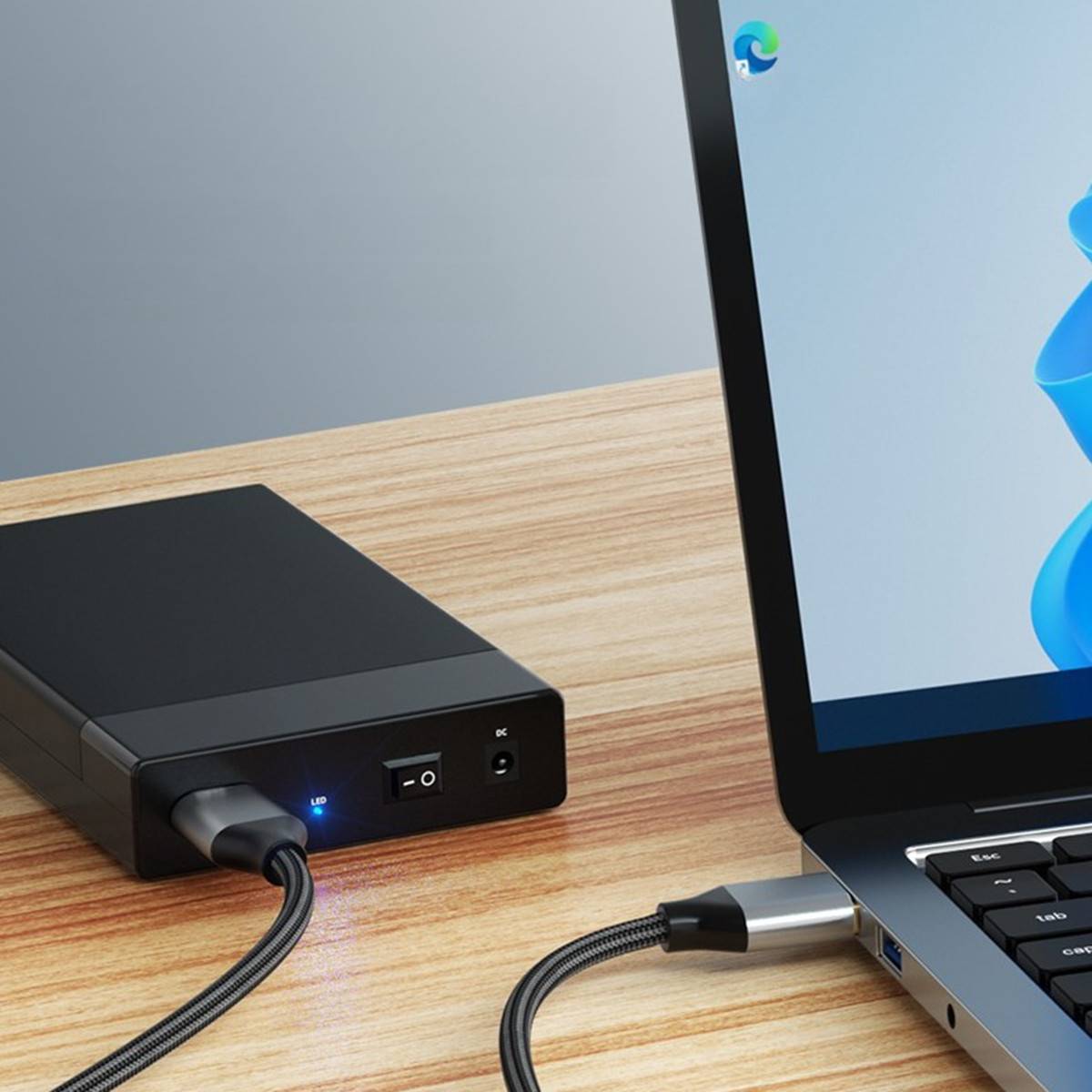 Laptop verbunden mit einer schwarzen externen Powerbank/Ladegerät auf einem Holzschreibtisch, mit Kabeln, die in beide Geräte eingesteckt sind.