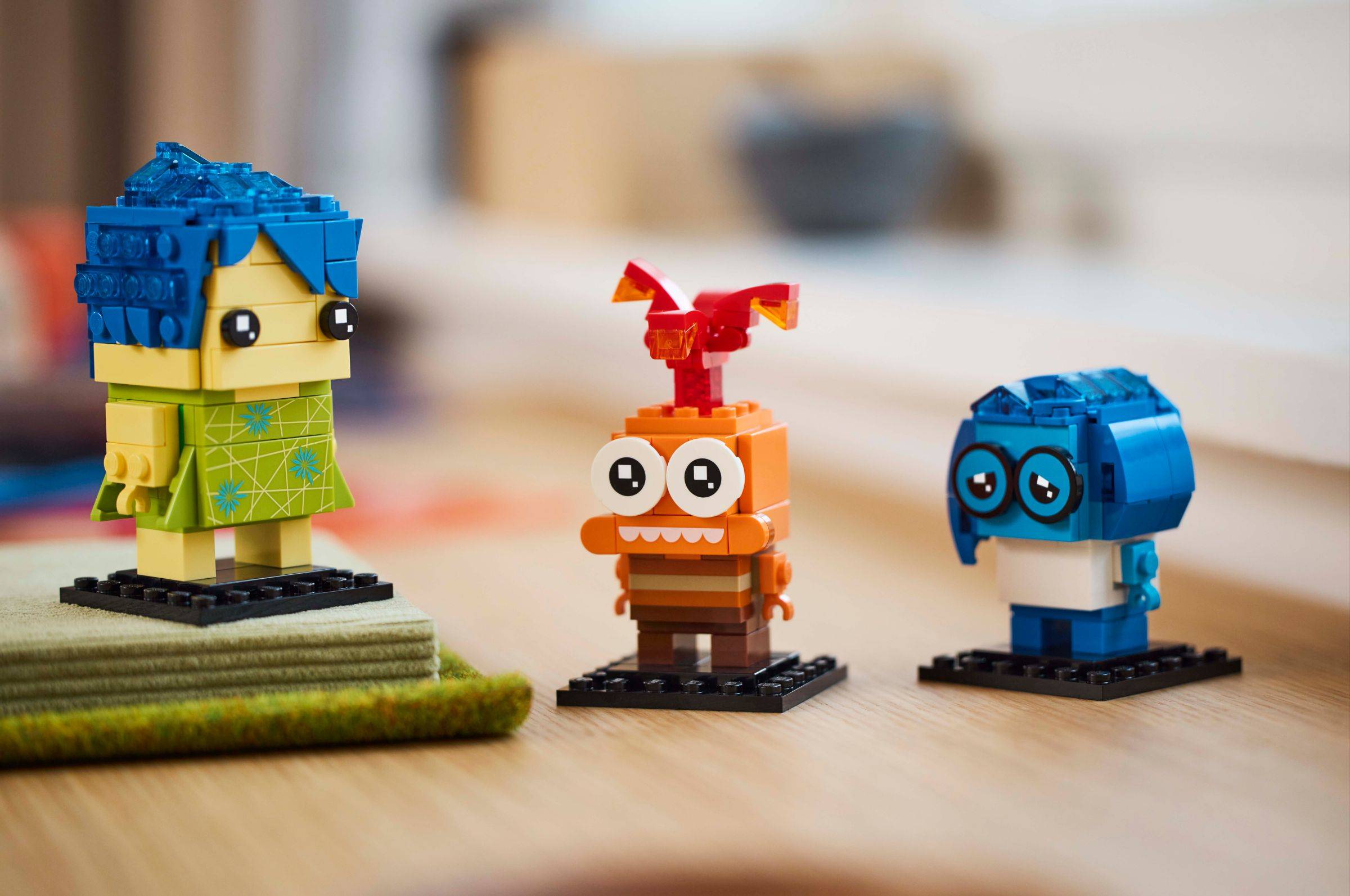 Drei kleine LEGO-ähnliche Charakterfiguren stehen auf einer Tischplatte, darunter eine blauharige Figur auf der linken Seite, eine orangefarbene Figur in der Mitte und eine blaue Figur mit Brille auf der rechten Seite.