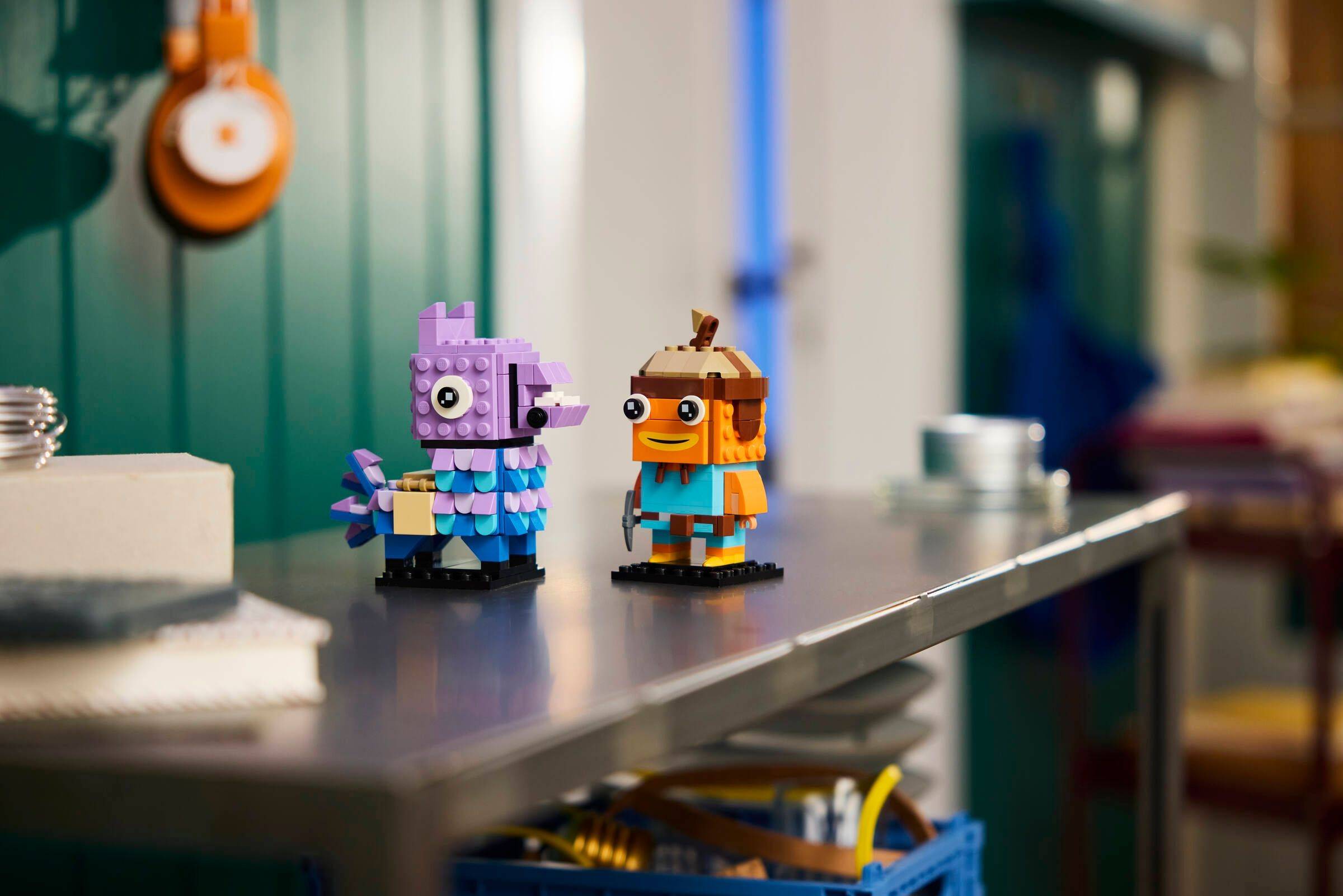 Zwei kleine LEGO-ähnliche Vogelfiguren, eine lila und eine orange, stehen auf einer Werkbank in einer Werkstatt.