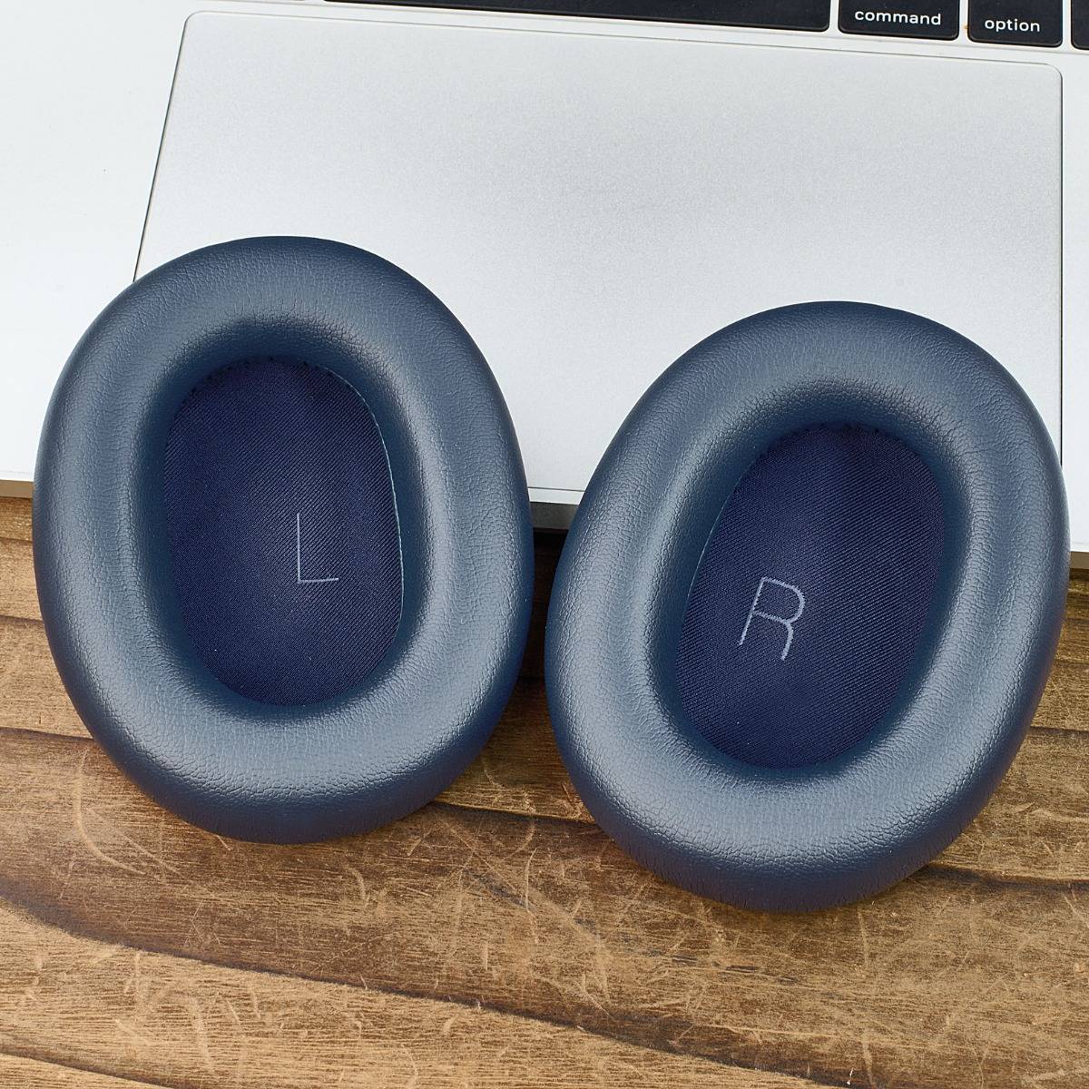 Paar blauer Over-Ear-Kopfhörer-Ohrpolster mit den Beschriftungen „L" und „R"", die vor einem Laptop auf einer Holzoberfläche ruhen.