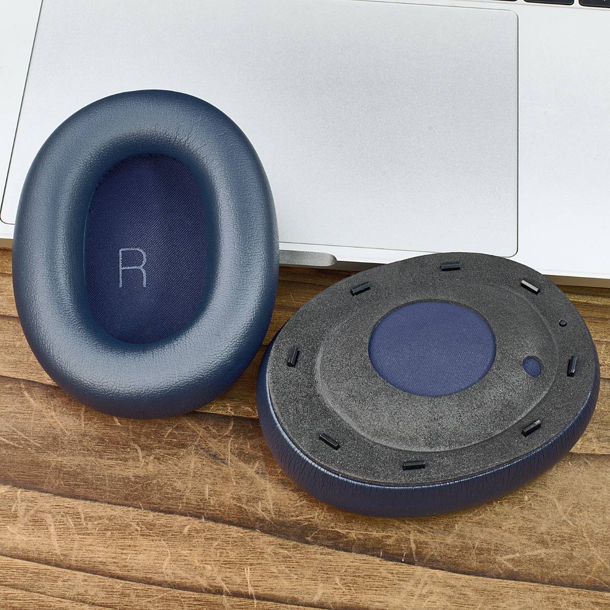 Zwei ovale blaue Ersatz-Ohrpolster für Kopfhörer, eines mit der Markierung „R" und eine Unteransicht, auf einer Holzoberfläche neben einem Laptop.