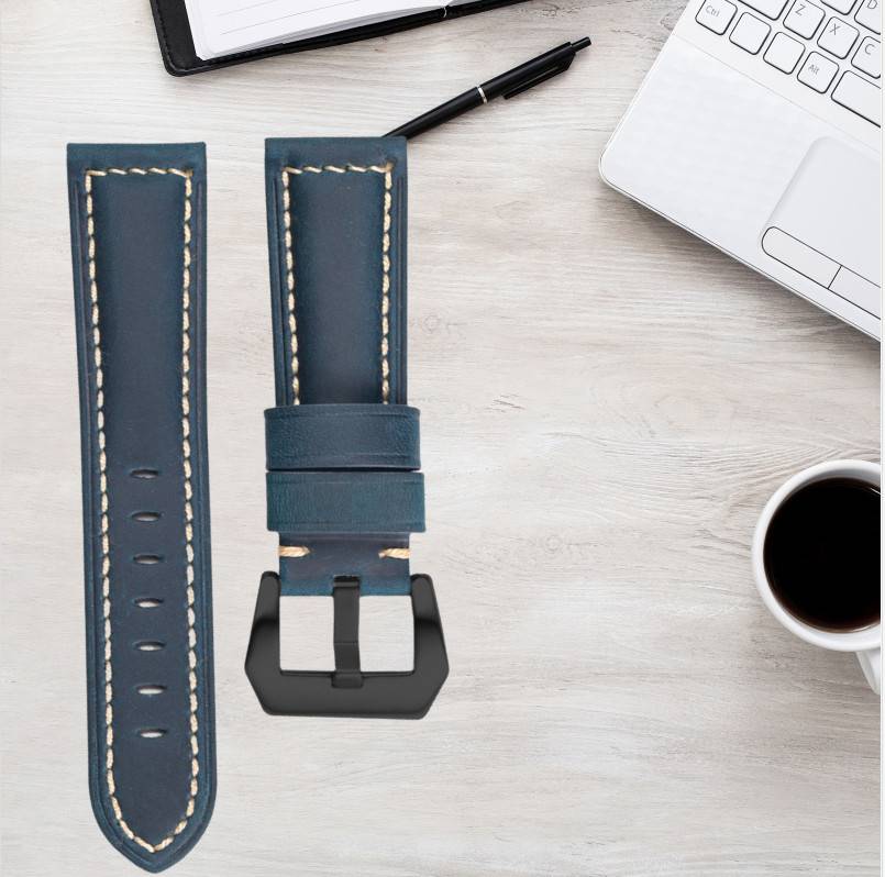 Blaues Lederarmband mit schwarzer Schließe auf einem Schreibtisch neben einem Laptop, Stift, Notizbuch und einer Kaffeetasse.