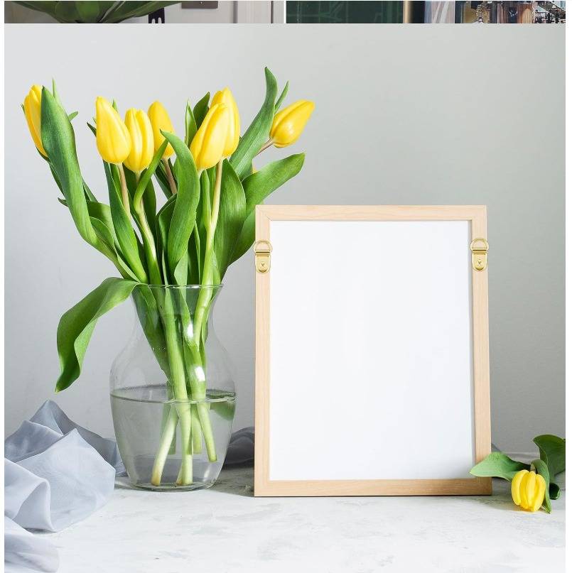 Gelbe Tulpen in einer Glasvase neben einem leeren Holzbilderrahmen auf einer weißen Oberfläche, mit einer lockeren Tulpe unten rechts.