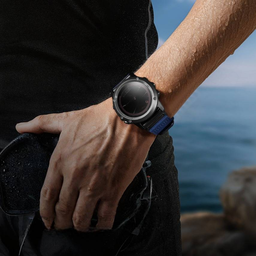 Nahaufnahme einer Hand einer Person, die eine runde schwarze Smartwatch mit blauem Armband am Meer trägt, was auf ein Outdoor-Abenteuer oder Fitness-Tracking hindeutet.