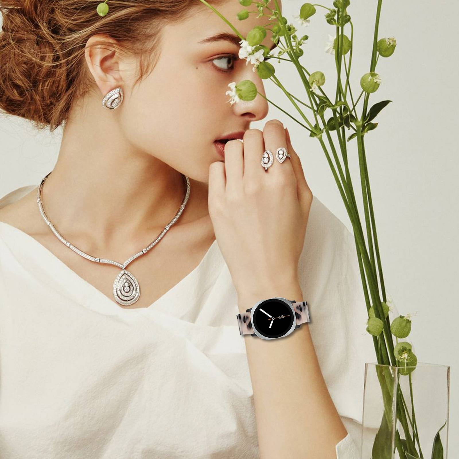 Frau in weißem Oberteil mit Diamantenschmuck und einer schwarzen Uhr, posierend neben grünen Blumen vor einem hellen Hintergrund.