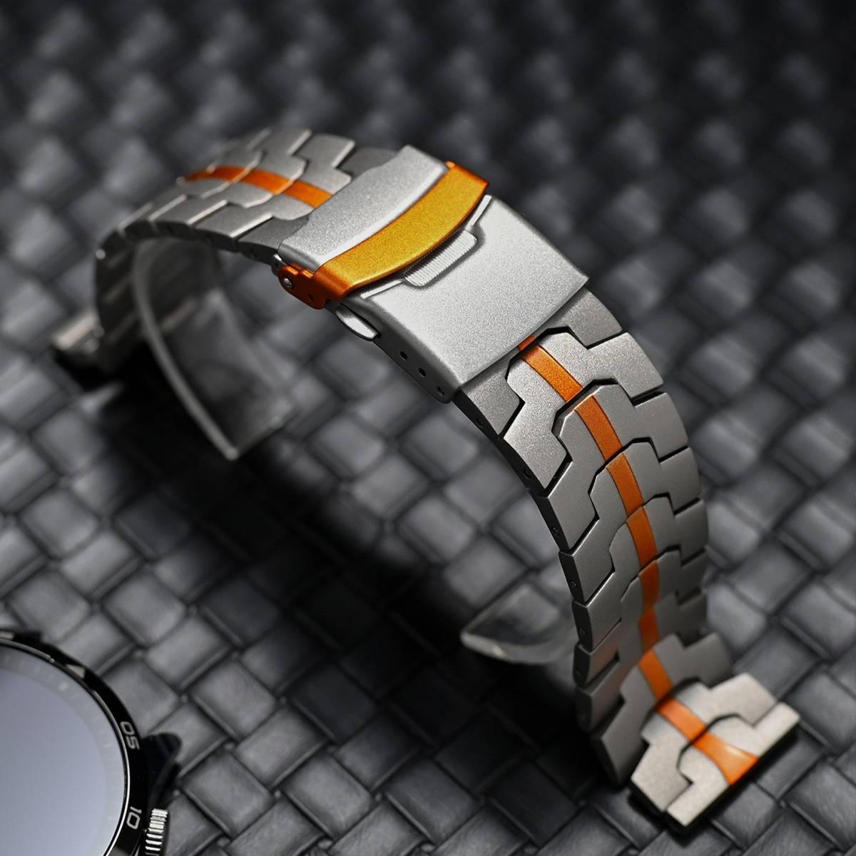 Nahaufnahme eines silbernen Metalluhrenarmband mit orangefarbenen Akzenten, auf einer dunklen gewebten Oberfläche gewölbt dargestellt.