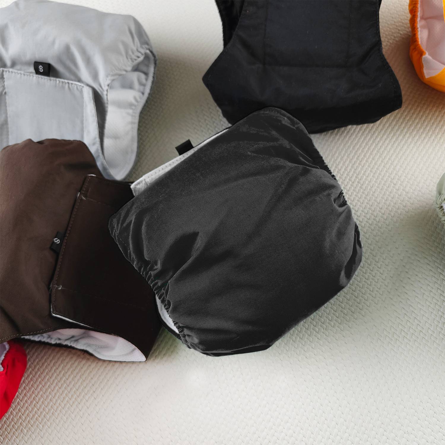 Mehrere gefaltete Stofftaschen oder Beutel in Grau, Schwarz, Braun und Orange liegen verstreut auf einer hellen strukturierten Oberfläche.