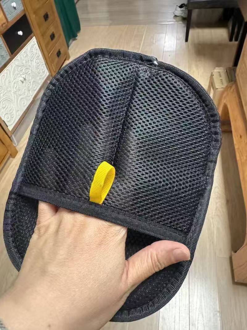 Eine Hand hält ein schwarzes Mesh-Oval-Pad oder eine schwarze Mesh-Tasche mit einer gelben Schlaufenlasche, dargestellt in Innenräumen auf einem Holzboden.