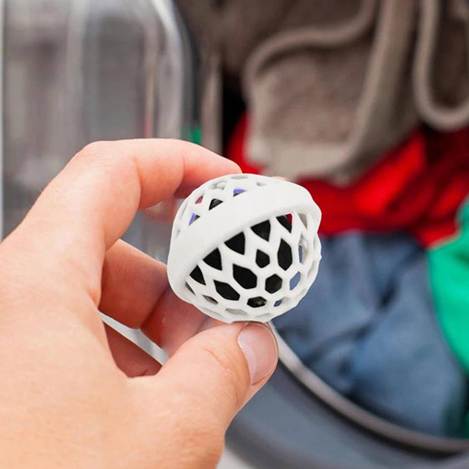 Hand, die einen kleinen weißen Gitterball hält, mit einem Wäschekorb und gefalteter Wäsche im Hintergrund.
