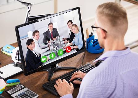 Ein Mann im Büro, der einen Computer für eine Videokonferenz mit vier Kollegen auf dem Monitor nutzt, wobei Anrufsteuerungen sichtbar sind.