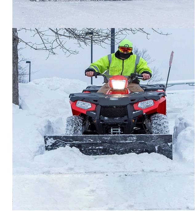 Eine Person in einer Neonjacke fährt ein rotes Quad mit einer Schneefräse durch tiefen Schnee und räumt einen verschneiten Weg frei.